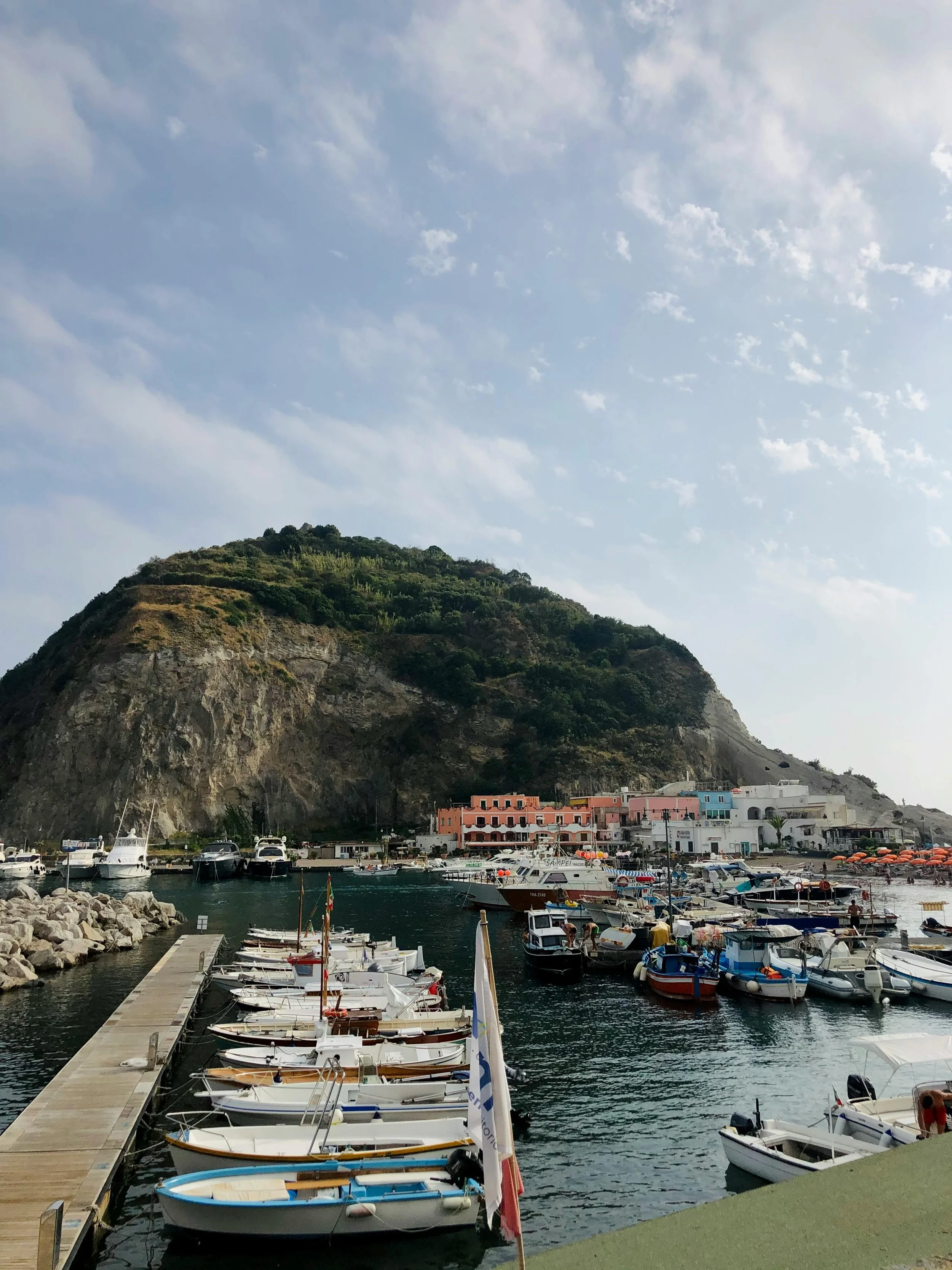 A harbor with boats docked along a pier, colorful buildings on a hillside, and a backdrop of a mountain under a partly cloudy sky.