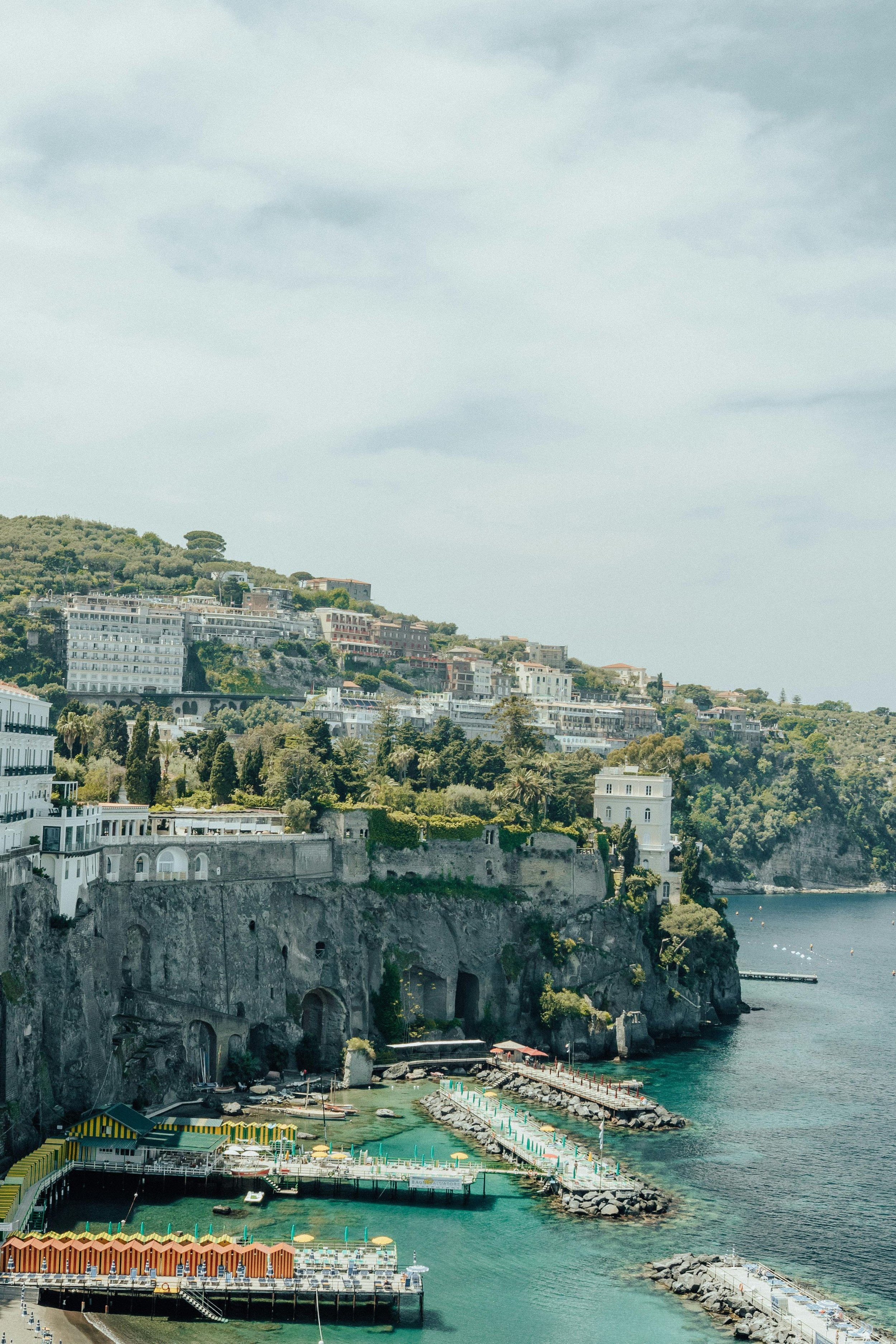 A coastal scene with buildings on a hillside, near a rocky shoreline and a harbor with colorful structures and boats. Amalfi Coast, Italy