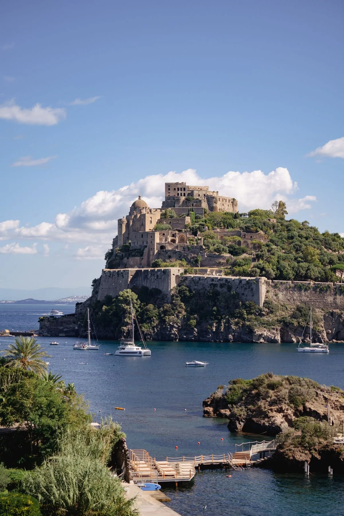 A historic fortress sits on top of a hill overlooking a harbor with boats and yachts, under a partly cloudy sky. Ischia italy
