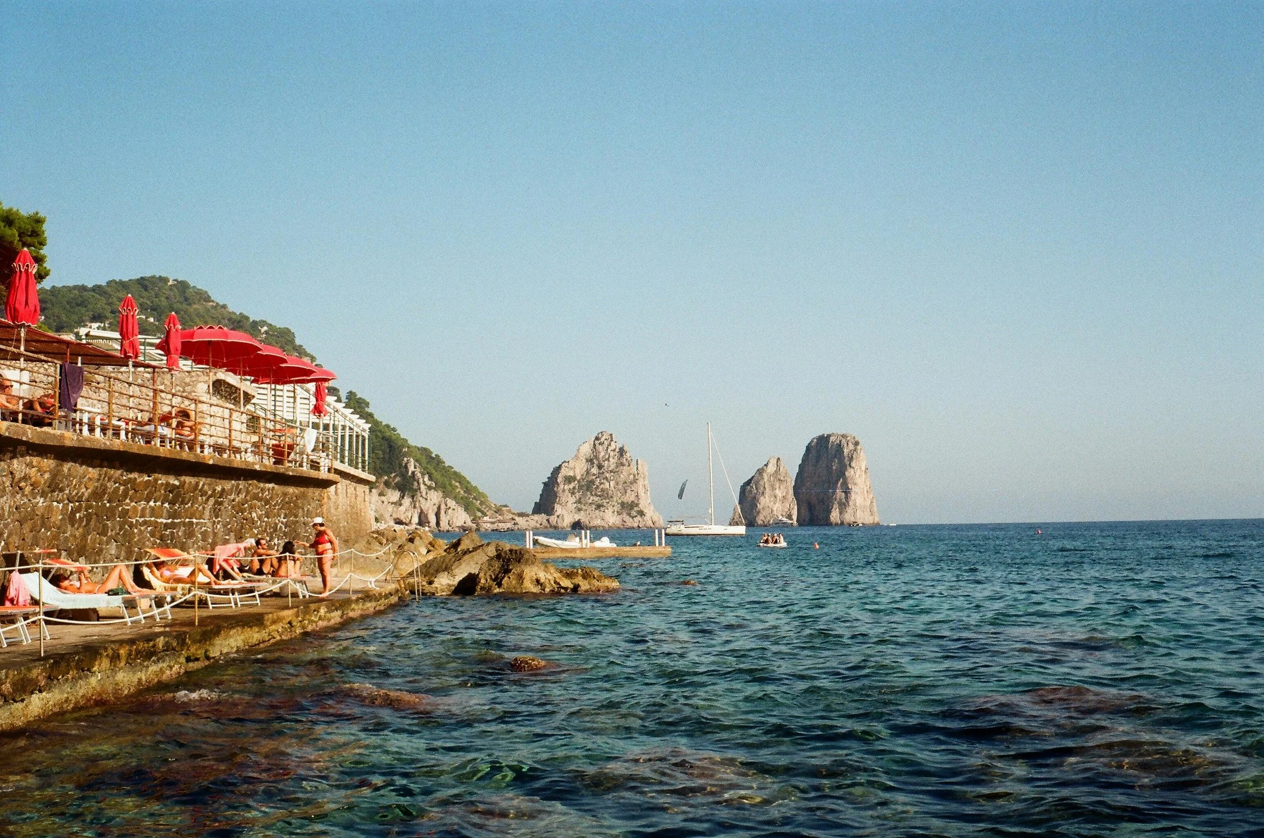 A seaside scene with people sunbathing on lounge chairs and umbrellas on a stone terrace, boats and large rock formations in the water, and a blue sky of Capri, Italy
