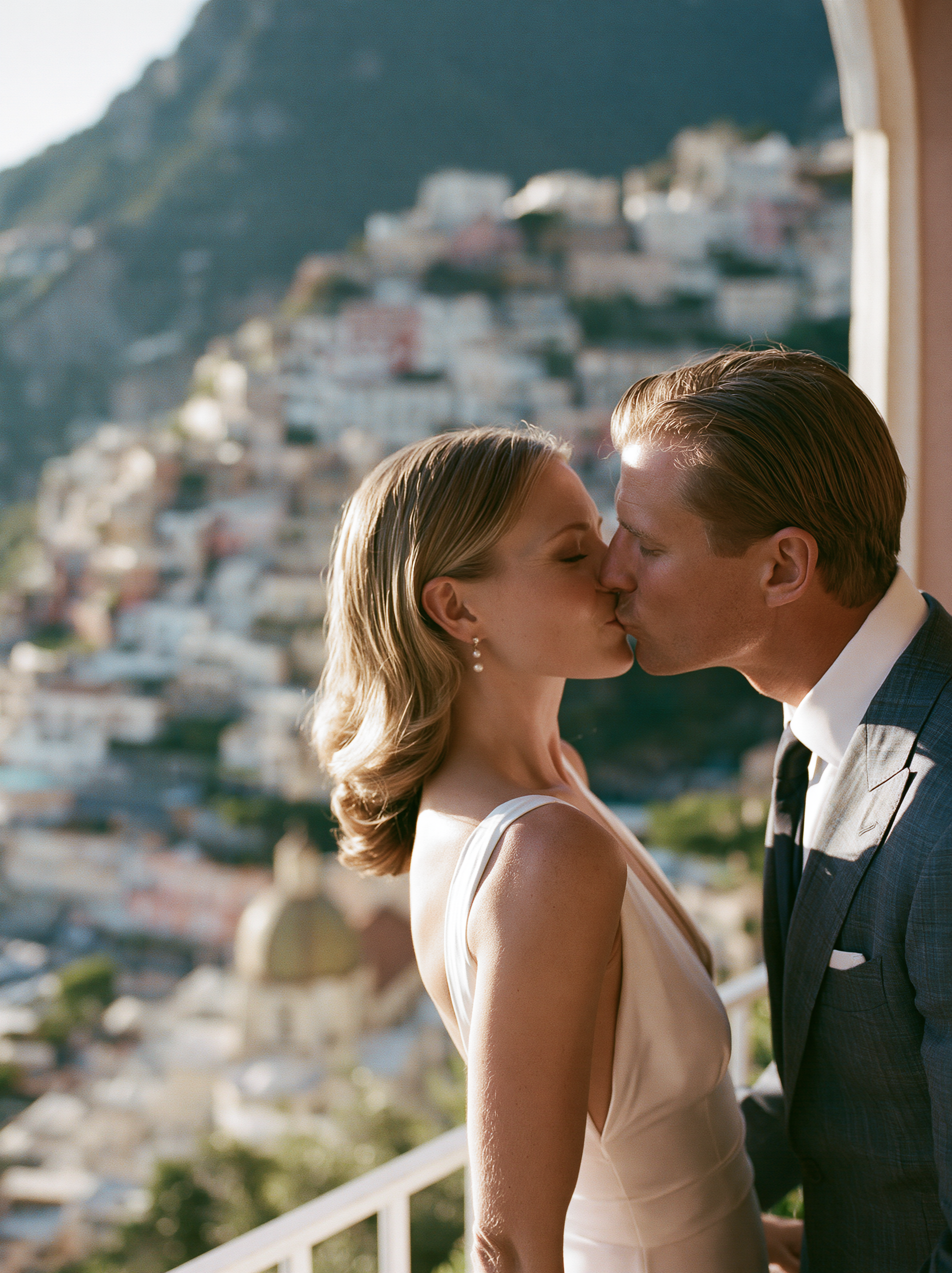 A man and woman kissing on a balcony overlooking a hillside town.