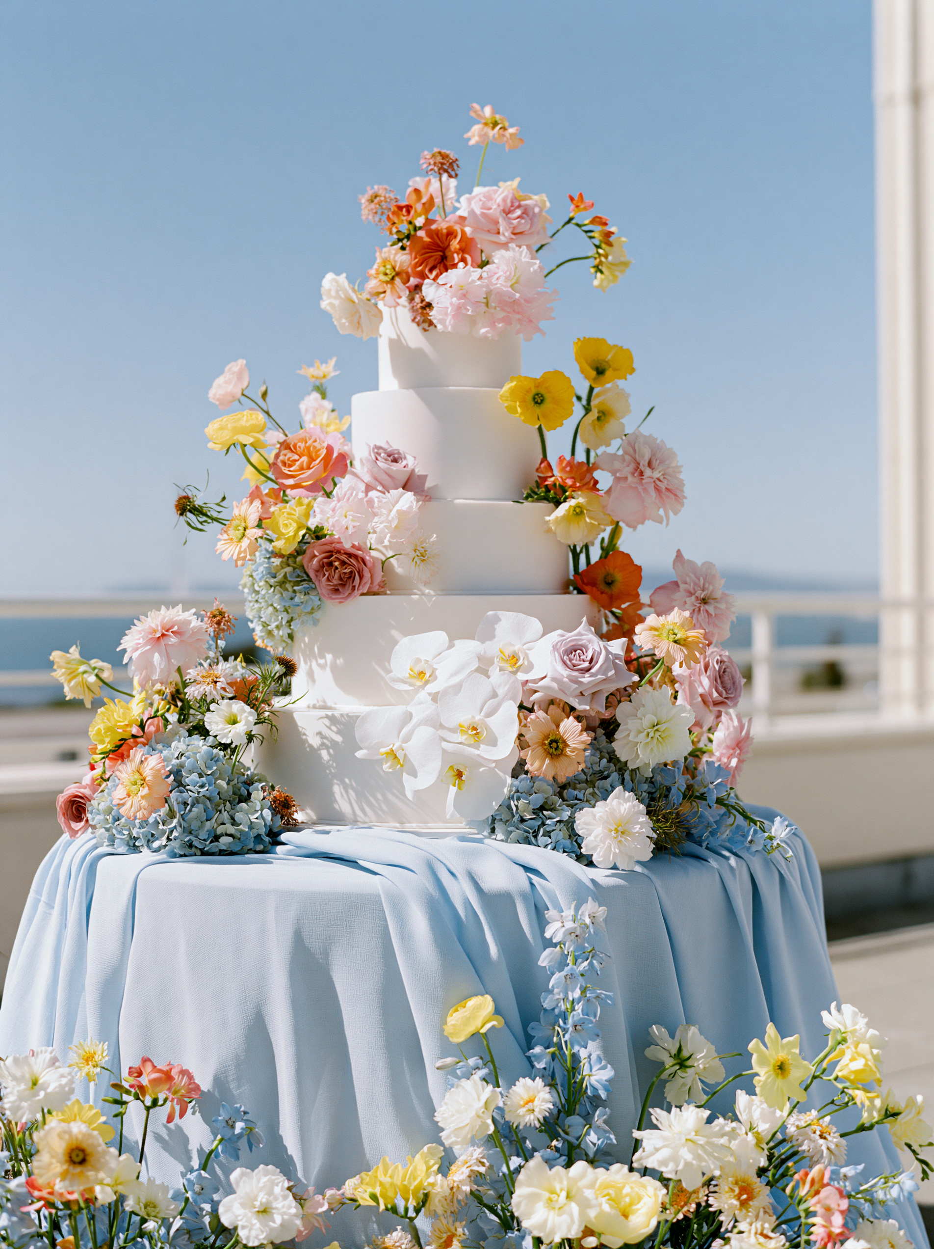 A tiered wedding cake decorated with pastel-colored flowers, placed on a table with a white tablecloth, outdoors with a blue sky in the background.