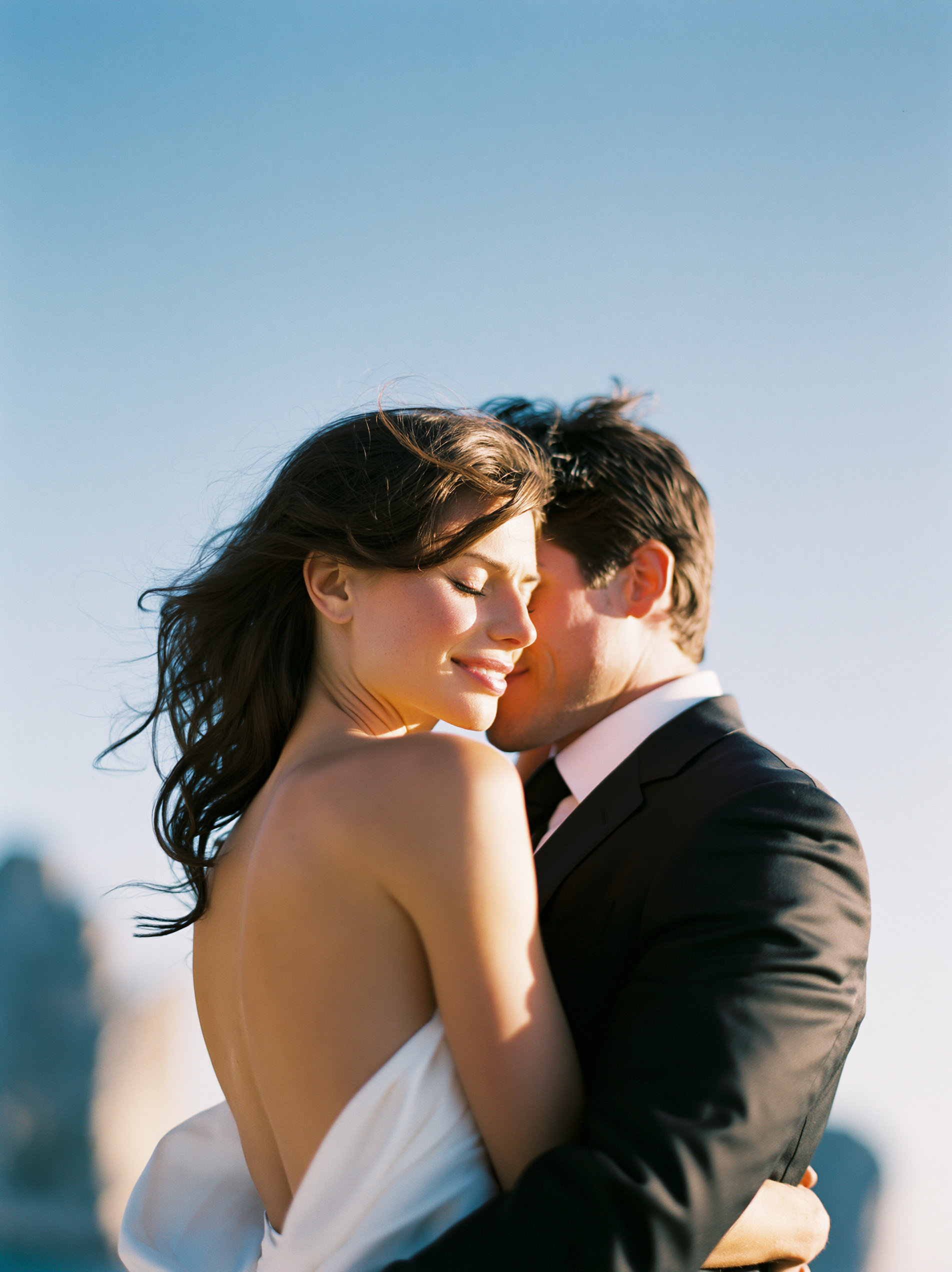 A romantic couple embracing outdoors during sunset, with the woman wearing a white dress and the man in a black suit, against a city skyline background.