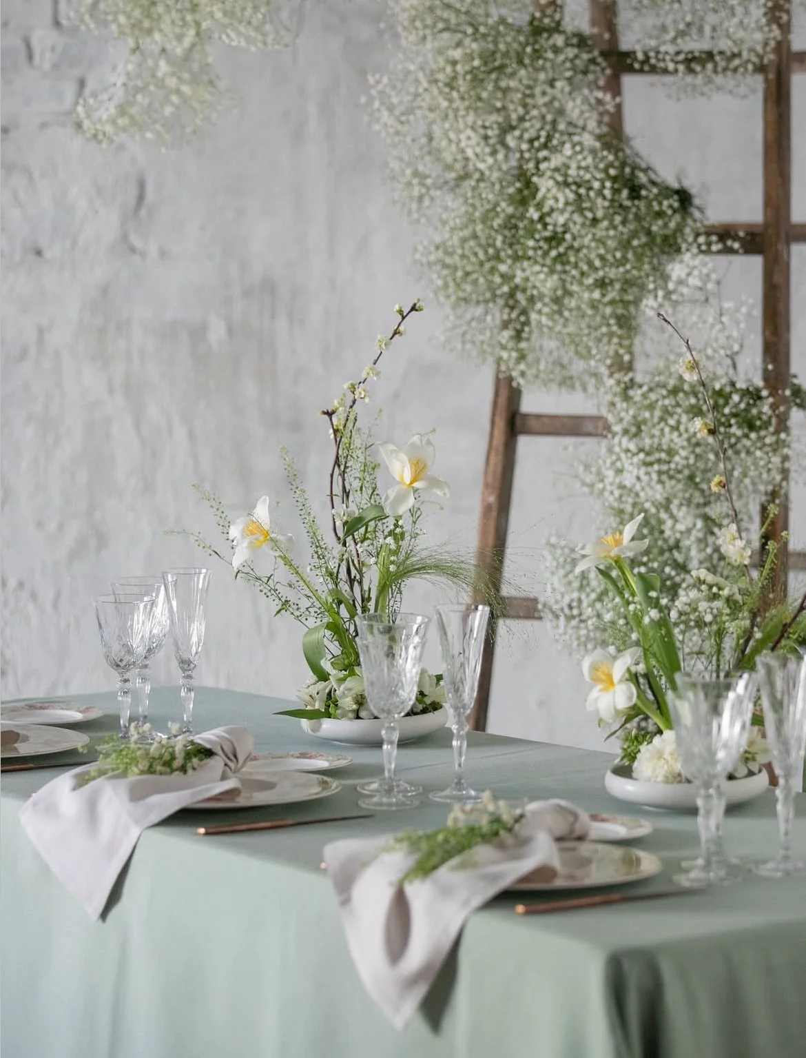 Table set with white plates, glassware, and white cloth napkins, decorated with white and green floral centerpieces on a green tablecloth, with a wooden ladder and greenery in the background.