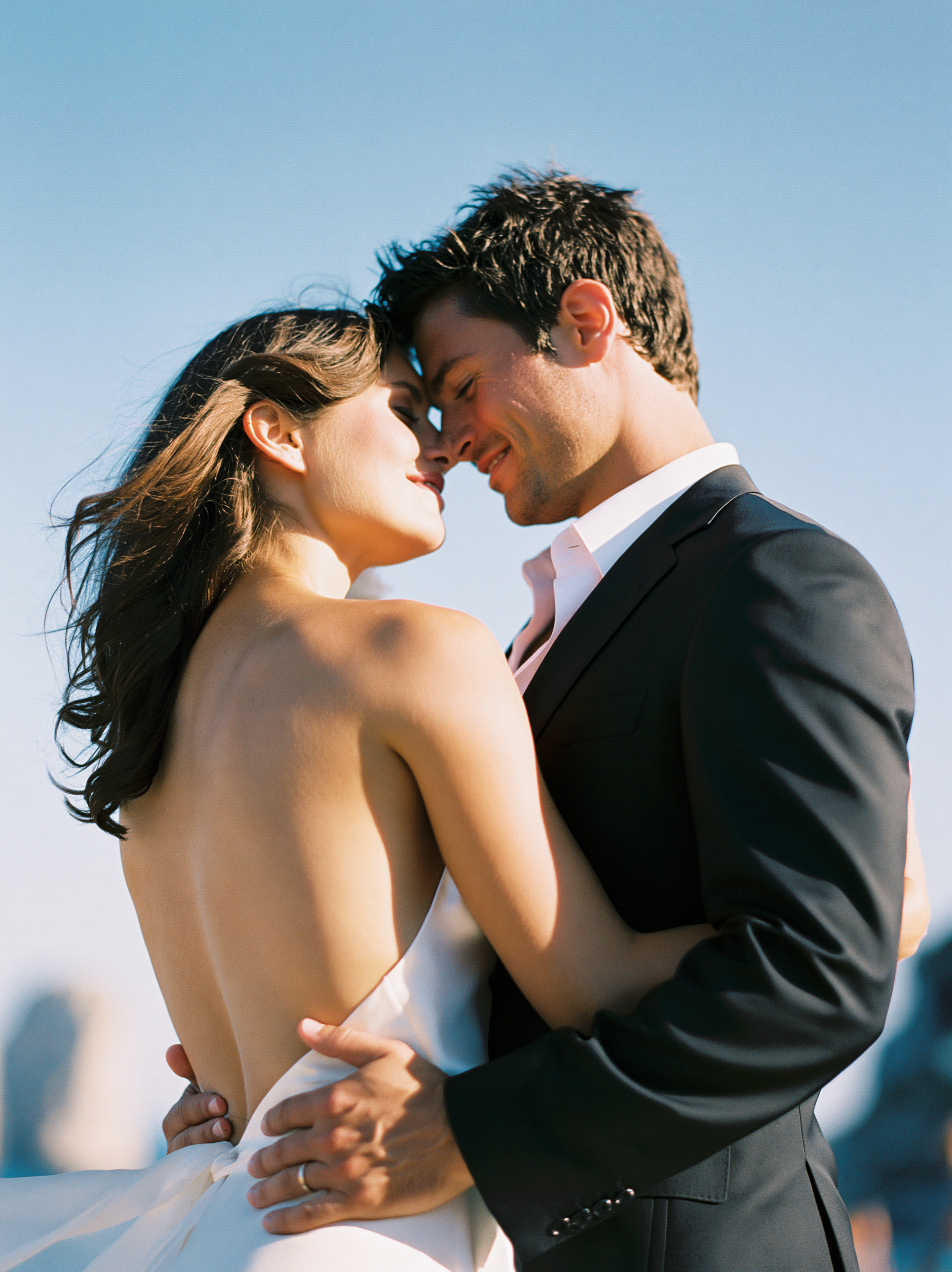 A couple dressed in wedding attire embracing outdoors during daytime, with clear blue sky in the background.