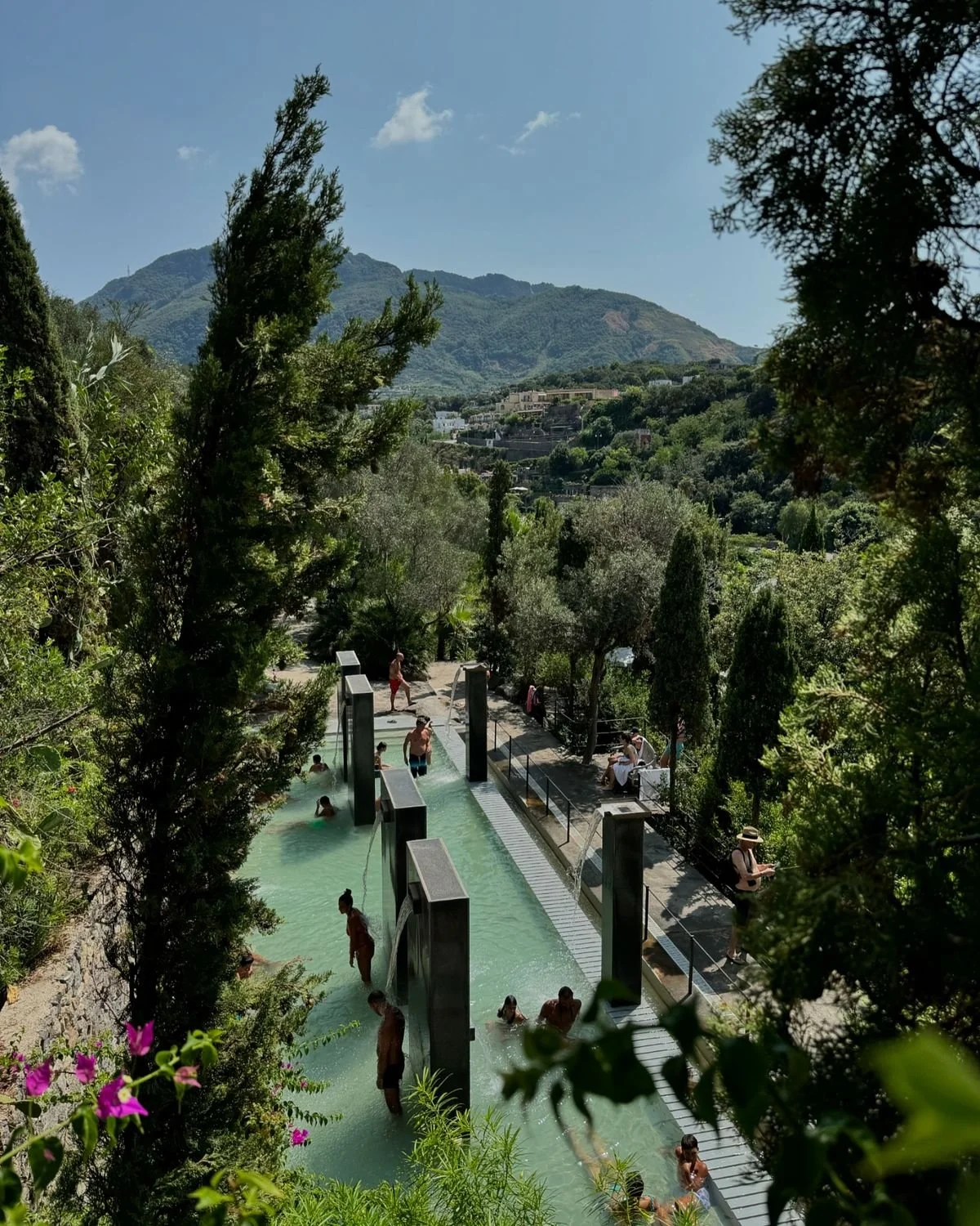 People enjoying a hot springs pool on a hillside surrounded by lush green trees and mountains in the background, under a blue sky with scattered clouds.