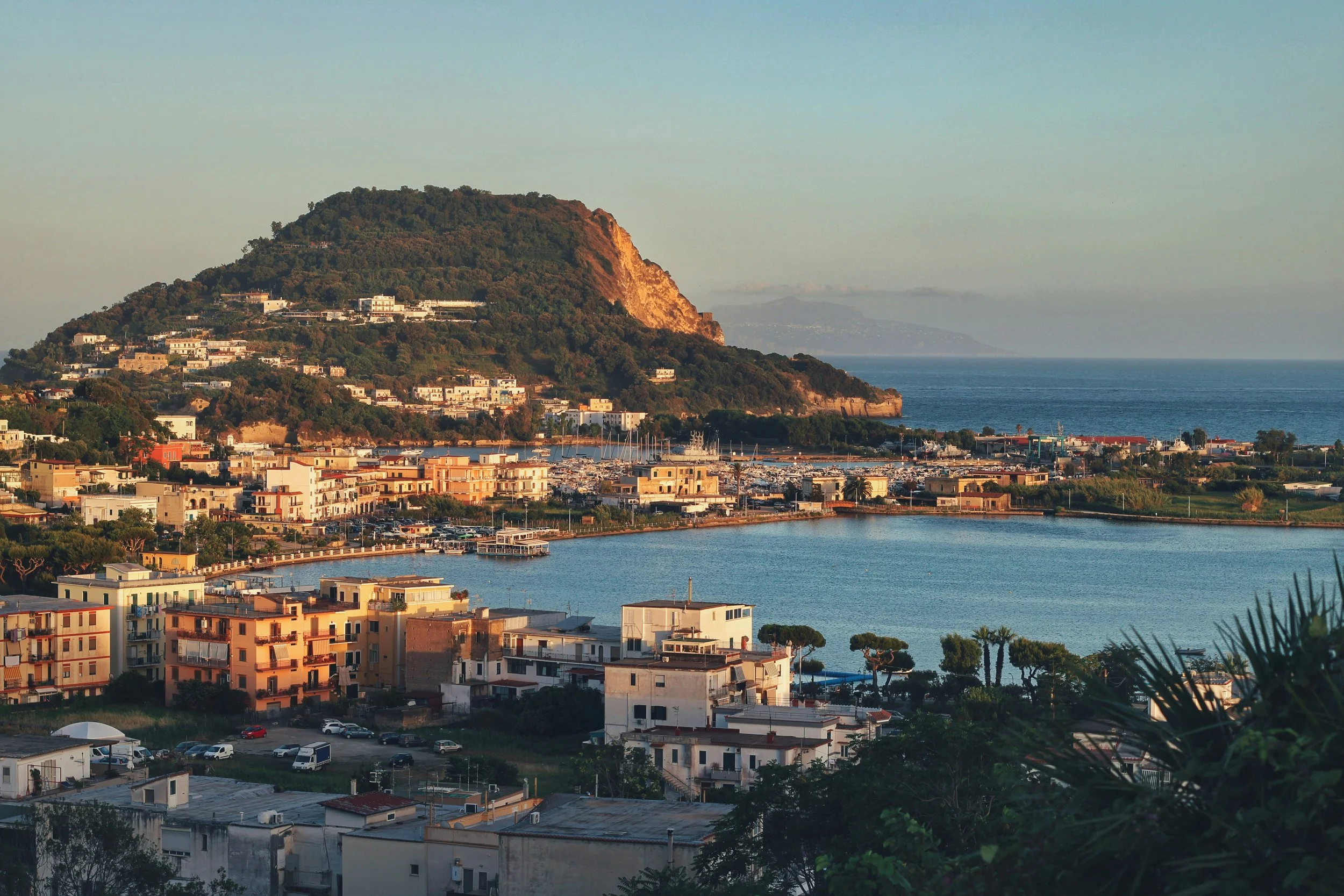Panoramica di una città costiera con edifici colorati, una baia e un monte verde sullo sfondo, al tramonto