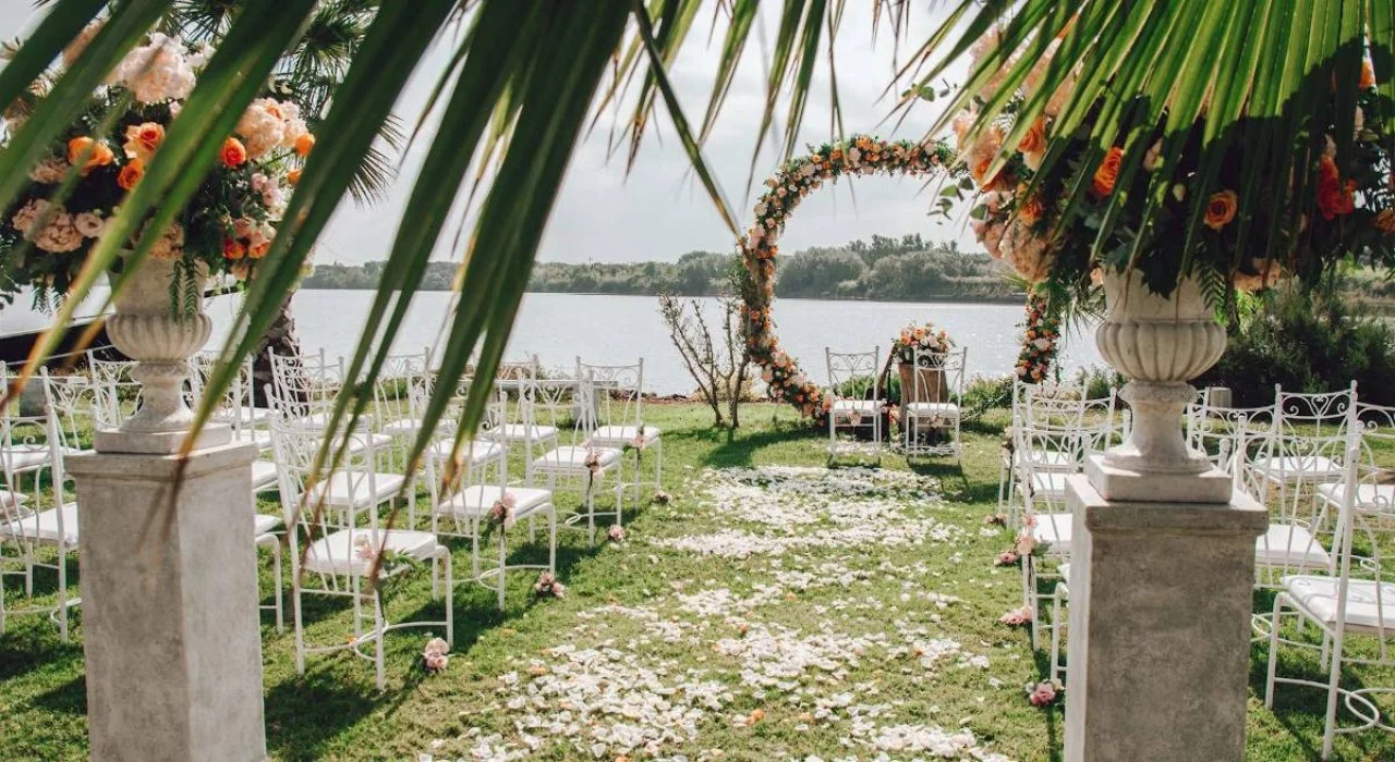 Sposalizio all'aperto sulla riva del lago con decorazioni floreali, sedie bianche e un arco di fiori colorati.