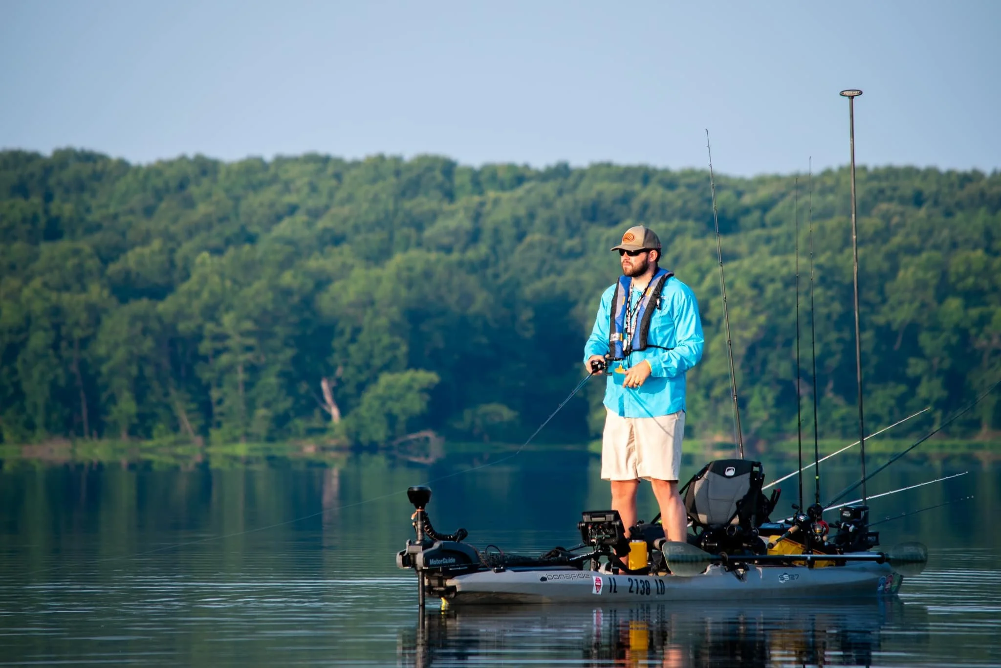 Man fishing on a small boat with fishing rods on calm lake with green forest in background.