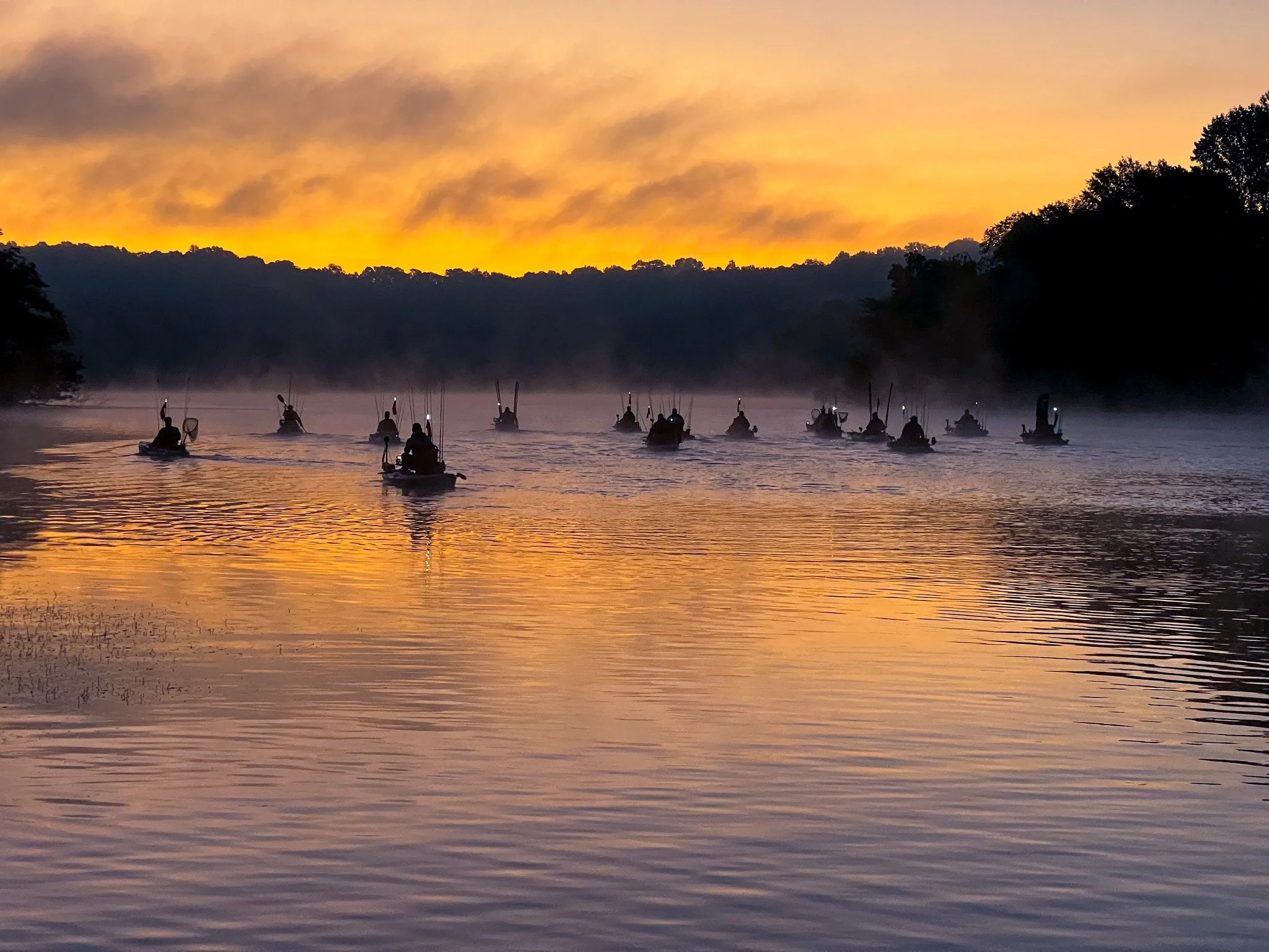Multiple silhouettes of people in small boats on a misty river at sunrise with a colorful sky and distant trees.