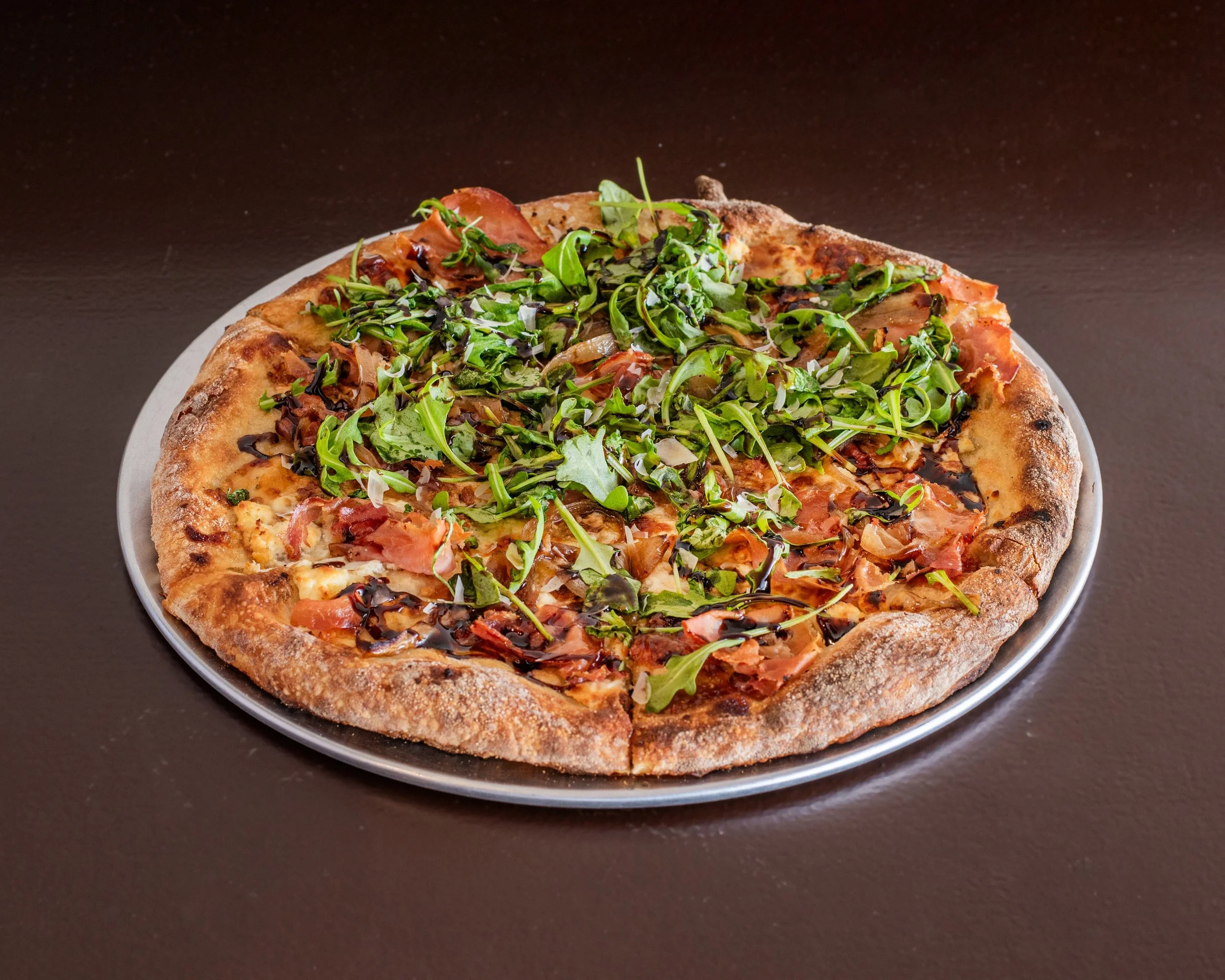 A pizza topped with fresh mixed greens and drizzled with balsamic glaze on a round metal tray