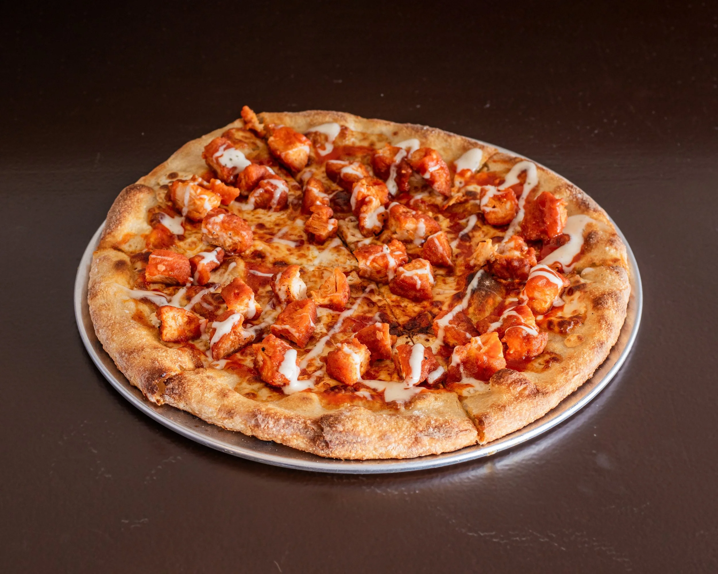 Pizza with chicken pieces, tomato sauce, melted cheese, and drizzled white sauce on a dark surface.