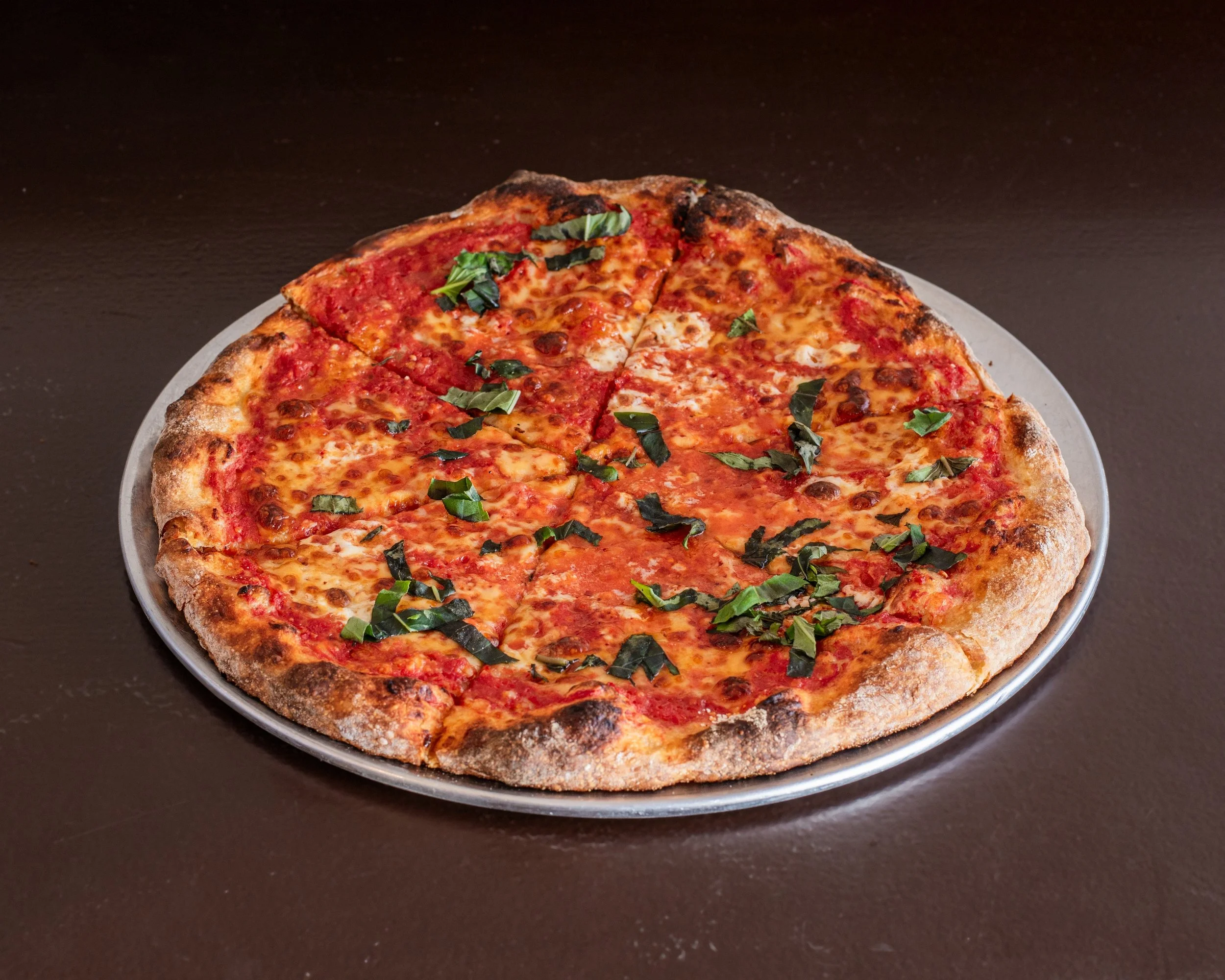 A pizza with a thin, crispy crust topped with tomato sauce, melted cheese, and fresh basil leaves, served on a round metal tray.