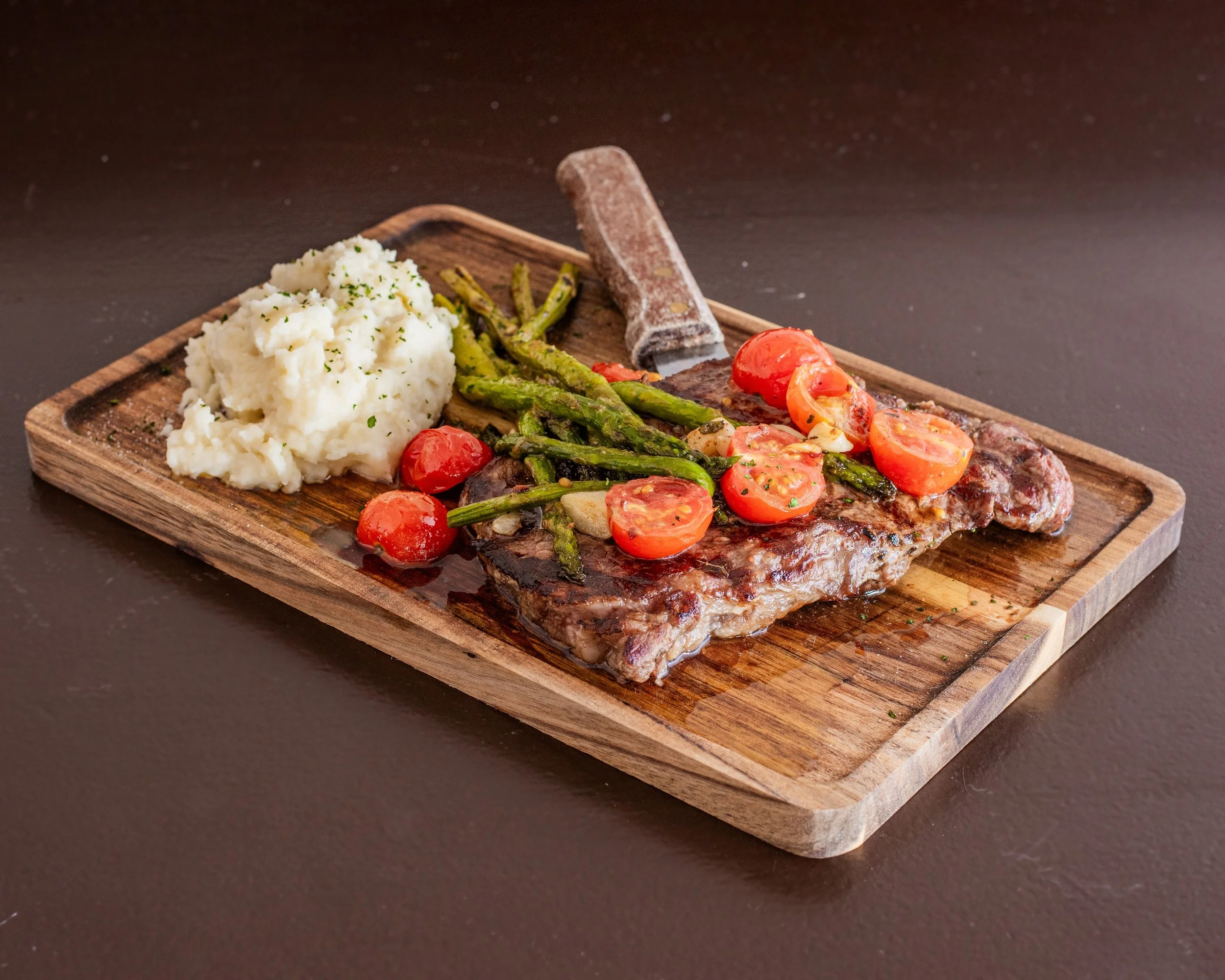 Grilled steak topped with cherry tomatoes and green beans on a wooden cutting board, with mashed potatoes on the side.