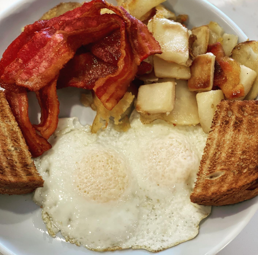 Breakfast plate with fried eggs, crispy bacon, home fries with cheese, and toasted bread.