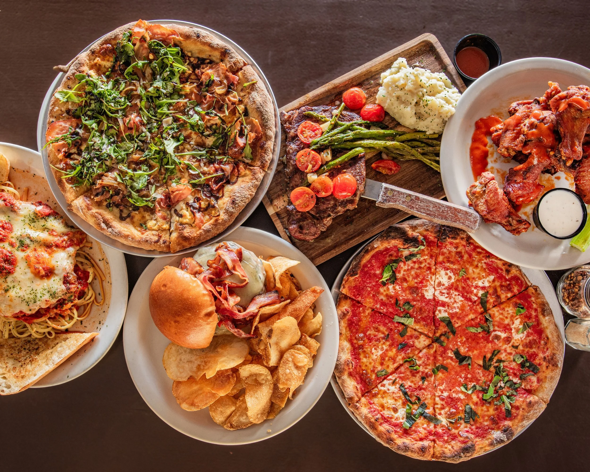 Assorted dishes including pizza, pasta, burger, chicken wings, and steak with sides on plates and a wooden serving board.