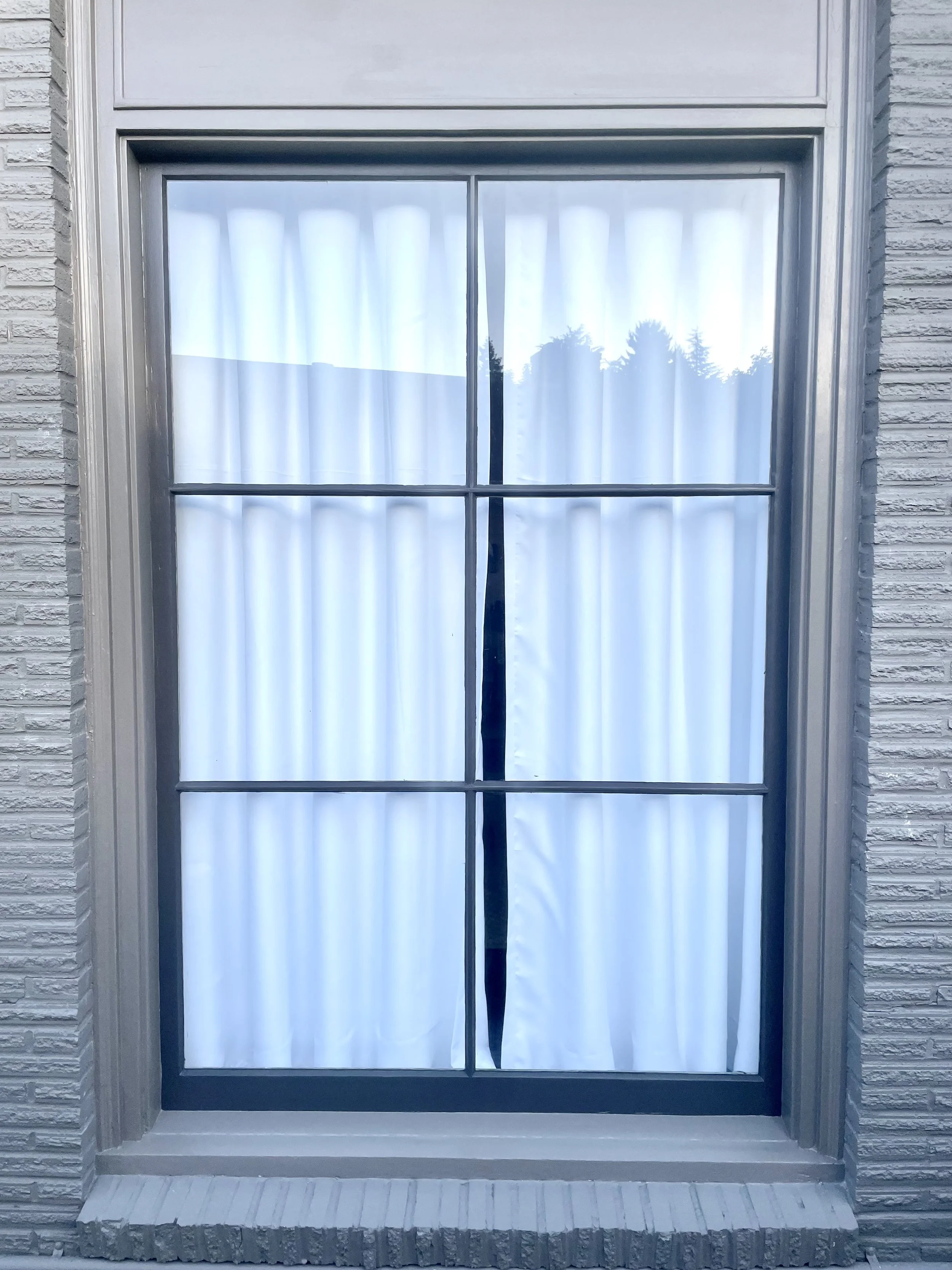 Close-up of a window with white curtains and a black tie-back, set in a brick wall.