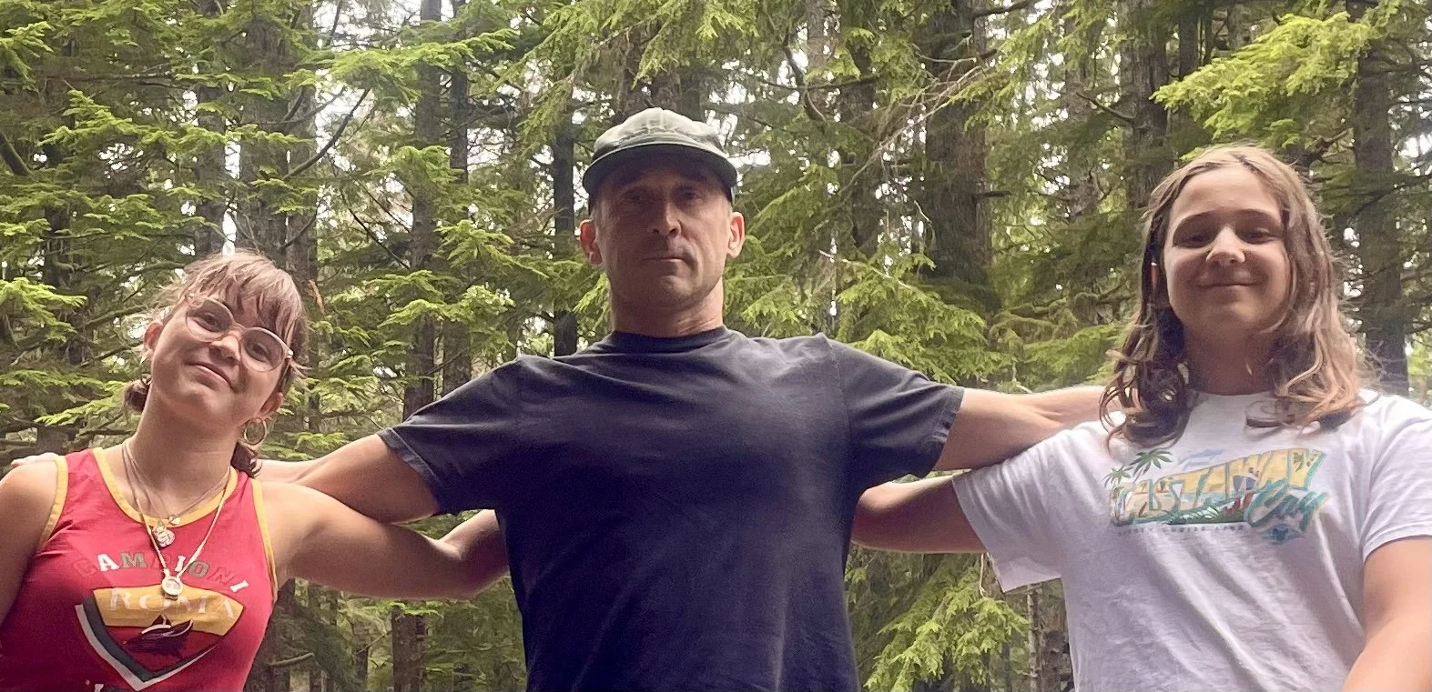 A man and two girls standing outdoors in a forest, with the man in the middle extending his arms around the girls, all smiling slightly.