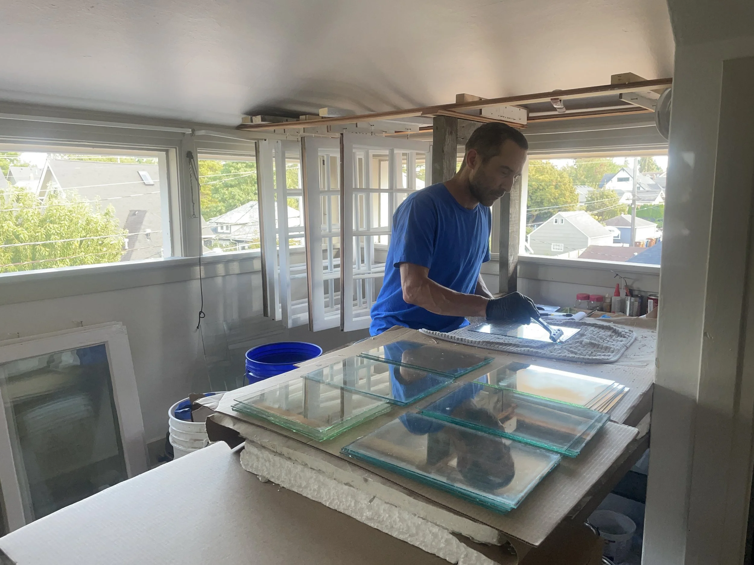A man in a blue shirt and gloves working on a stained glass project at a worktable inside a bright room with large windows showing a neighborhood view.