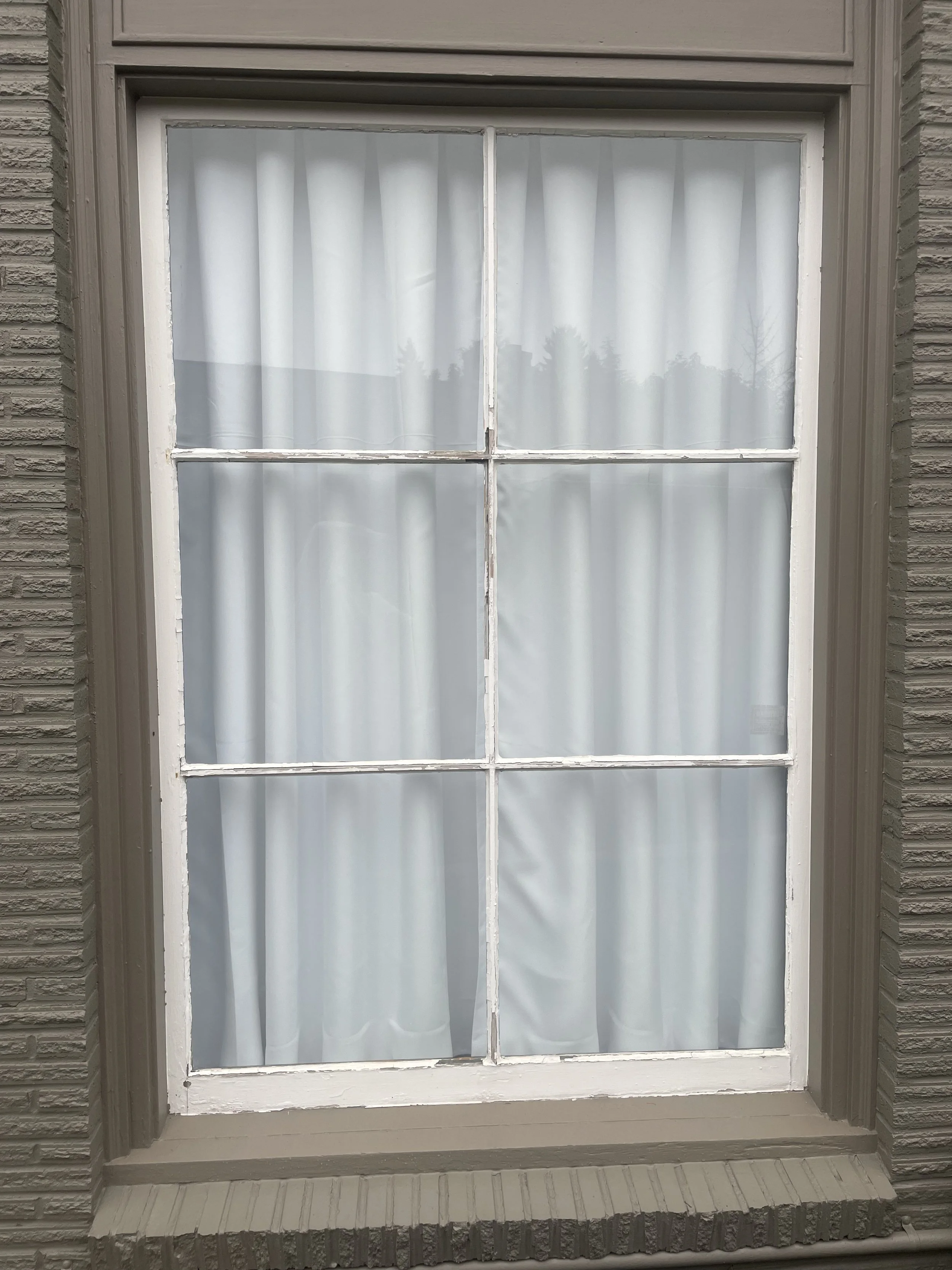 A wood window with white curtains and a white window frame. The window is divided into six panes, with side brick walls and a top trim around the window.