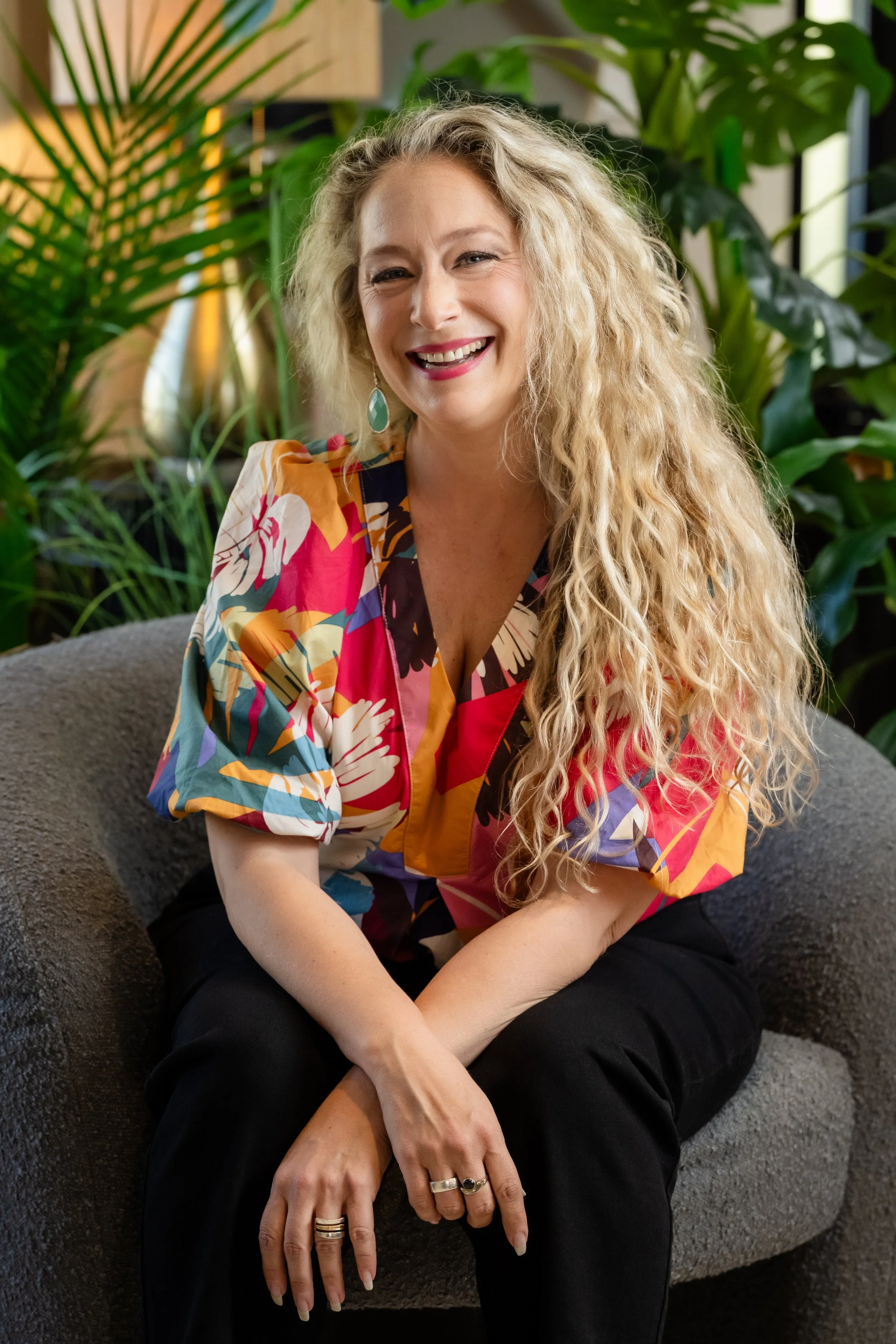 A woman with long, curly blonde hair smiling and sitting on a gray chair, surrounded by green plants in an indoor setting.