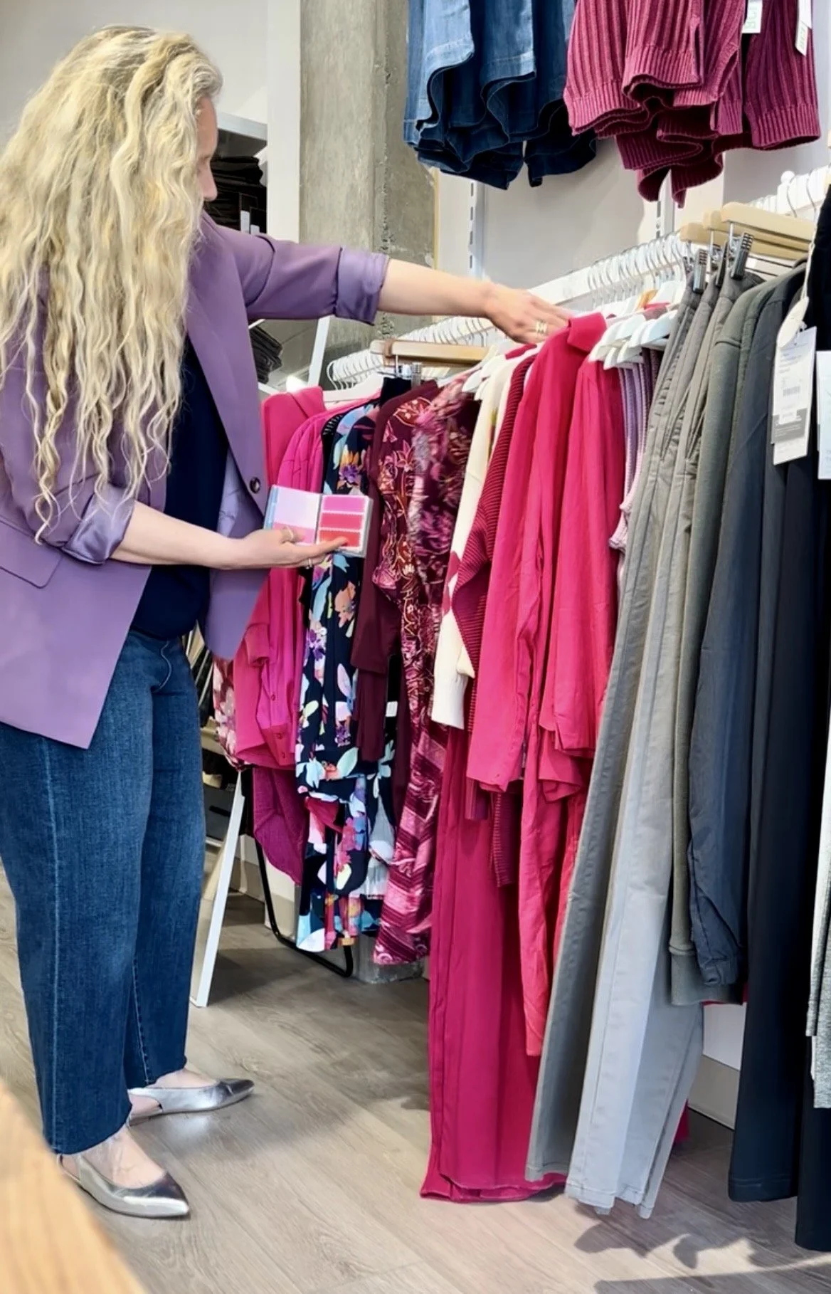 A woman shopping for clothes on a rack, holding a color palette, with various pink and floral garments around her.