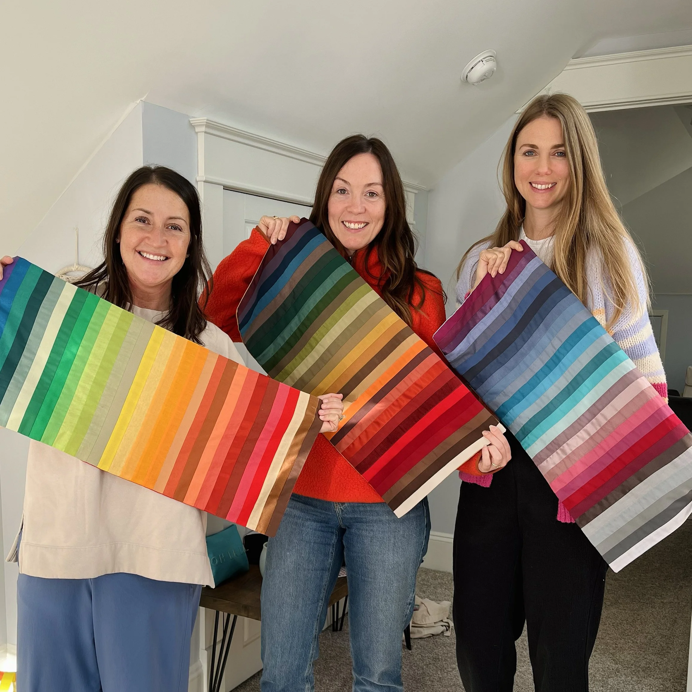 Three women standing indoors, holding large fabric swatches with colorful stripe patterns, smiling at the camera.