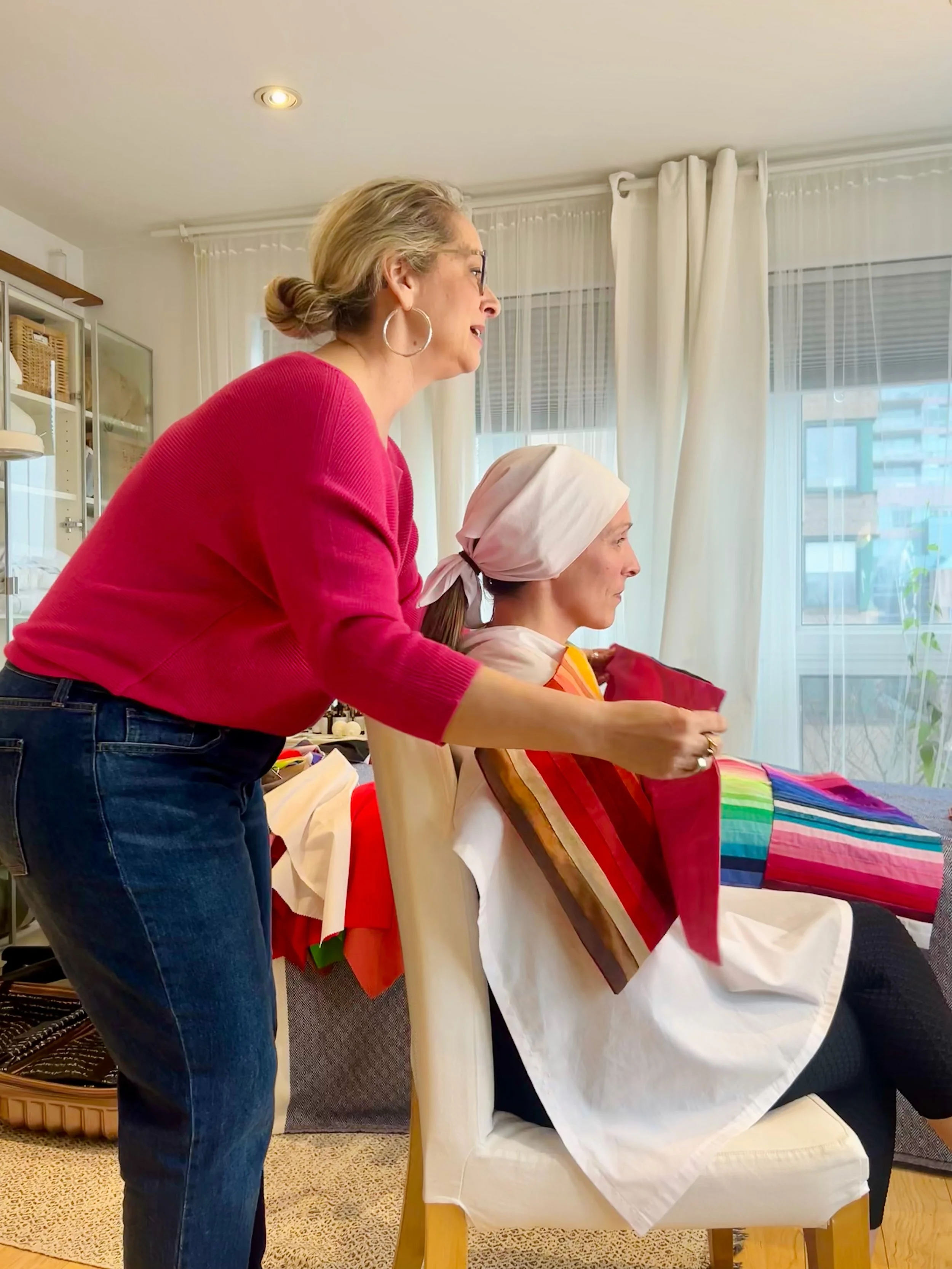 Two women, one sitting and one standing, in a brightly lit room, with the standing woman showing colorful fabric swatches to the seated woman.
