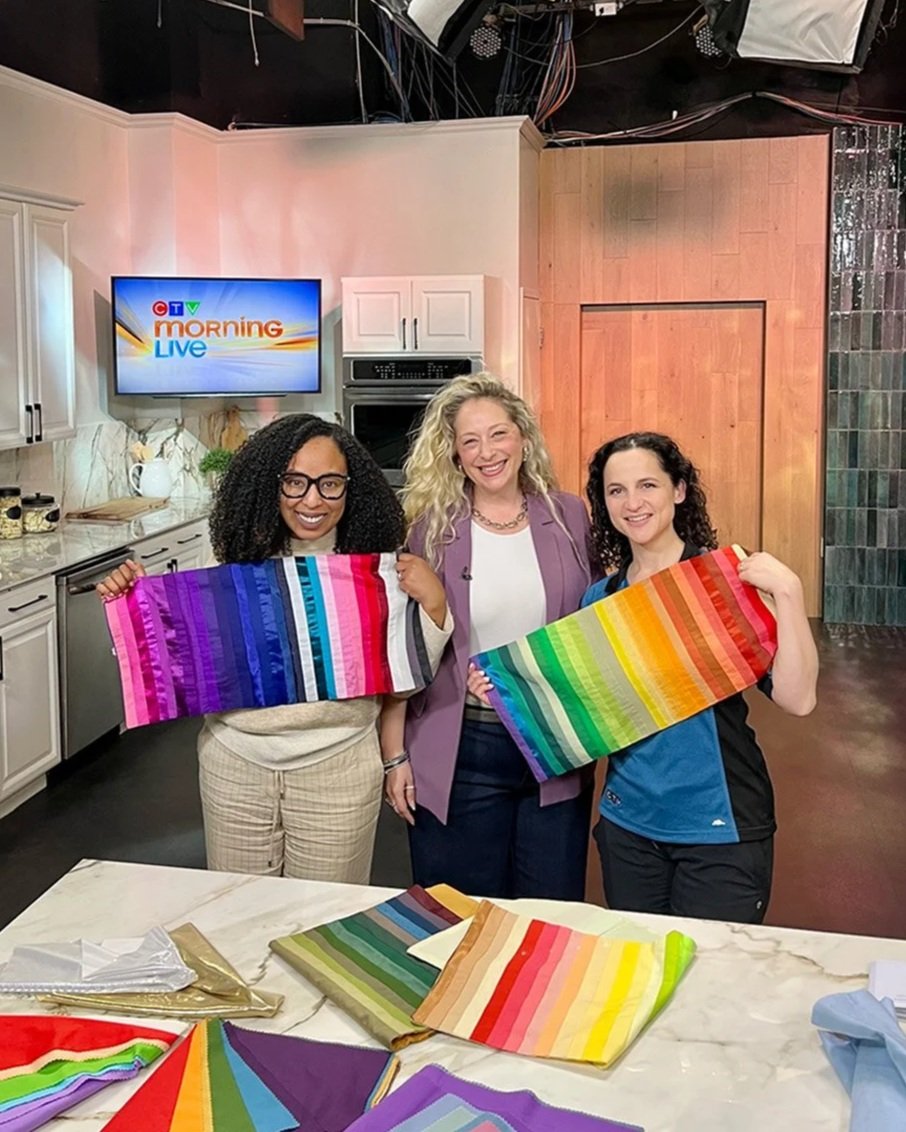 Three women standing in a kitchen holding colorful striped fabric, with more fabric laying on the table in front of them, smiling for the camera.
