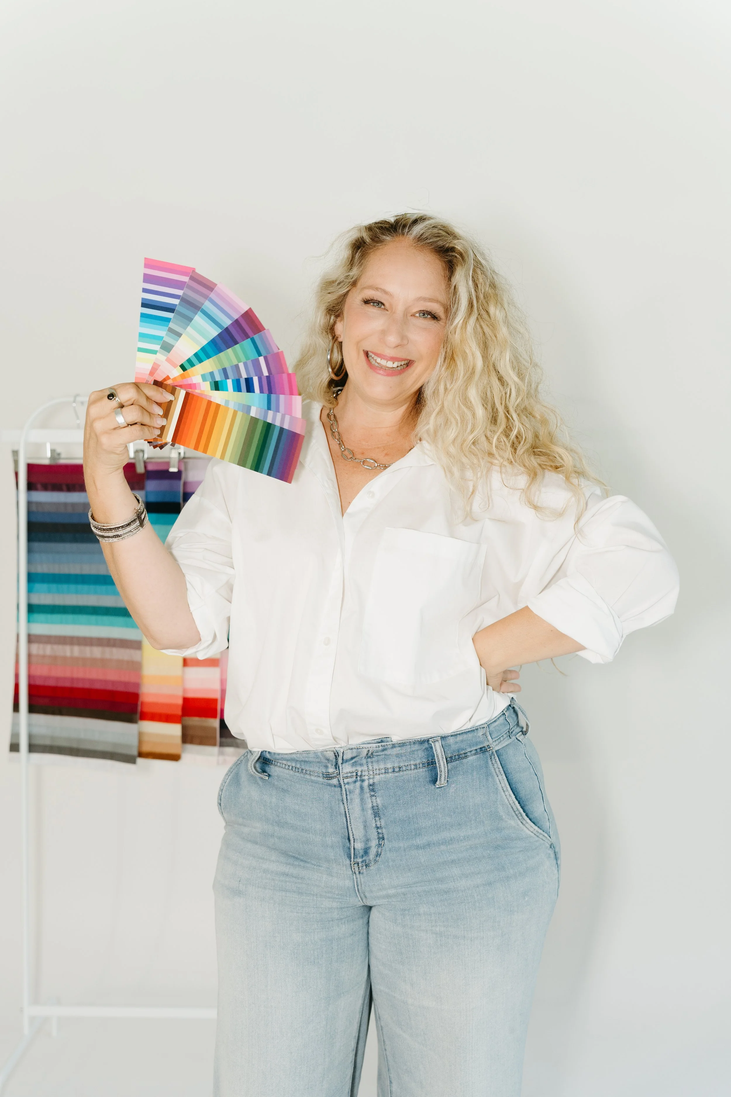 A smiling woman with curly blonde hair holding a fan of colorful paint swatches in a design studio.