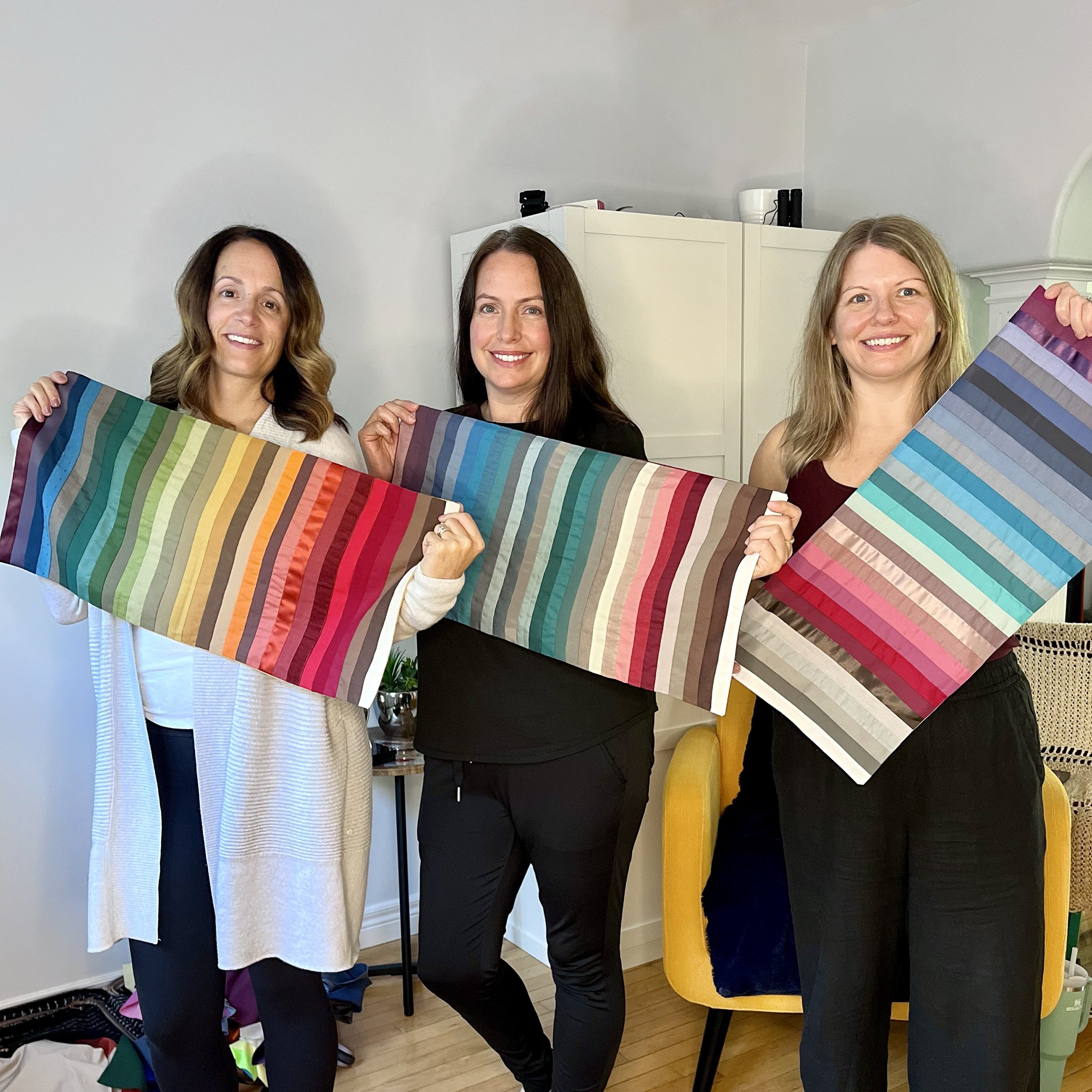 Three women standing indoors, each holding a colorful striped fabric or art piece, smiling at the camera.