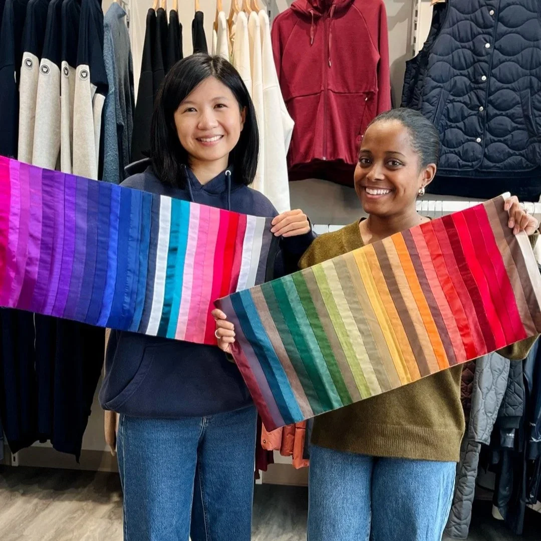 Two women smiling and holding colorful fabric swatches inside a clothing store with jackets hanging in the background.