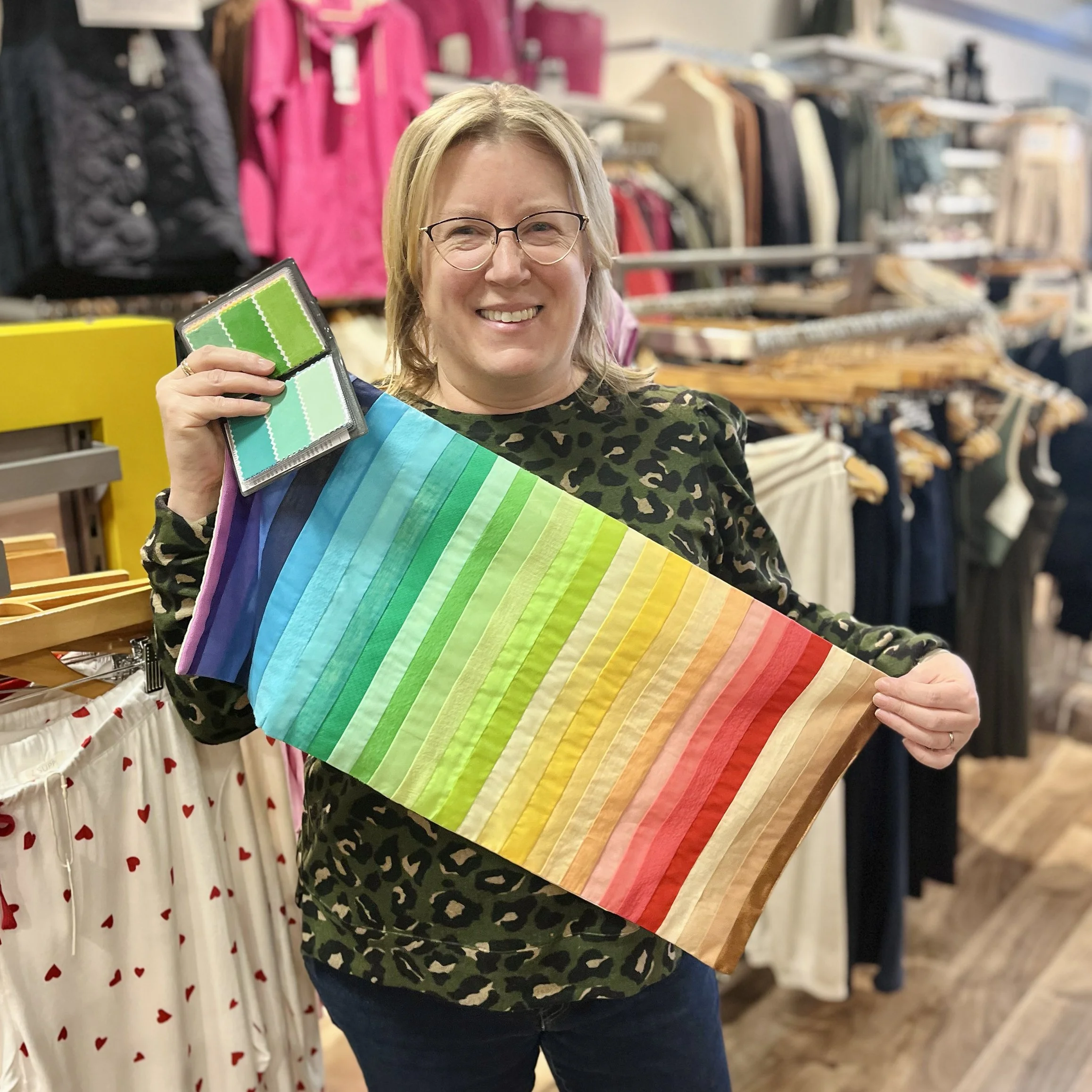 A woman in a leopard print shirt holding a rainbow-colored fabric sample and a smaller fabric swatch in a store with clothing racks in the background.