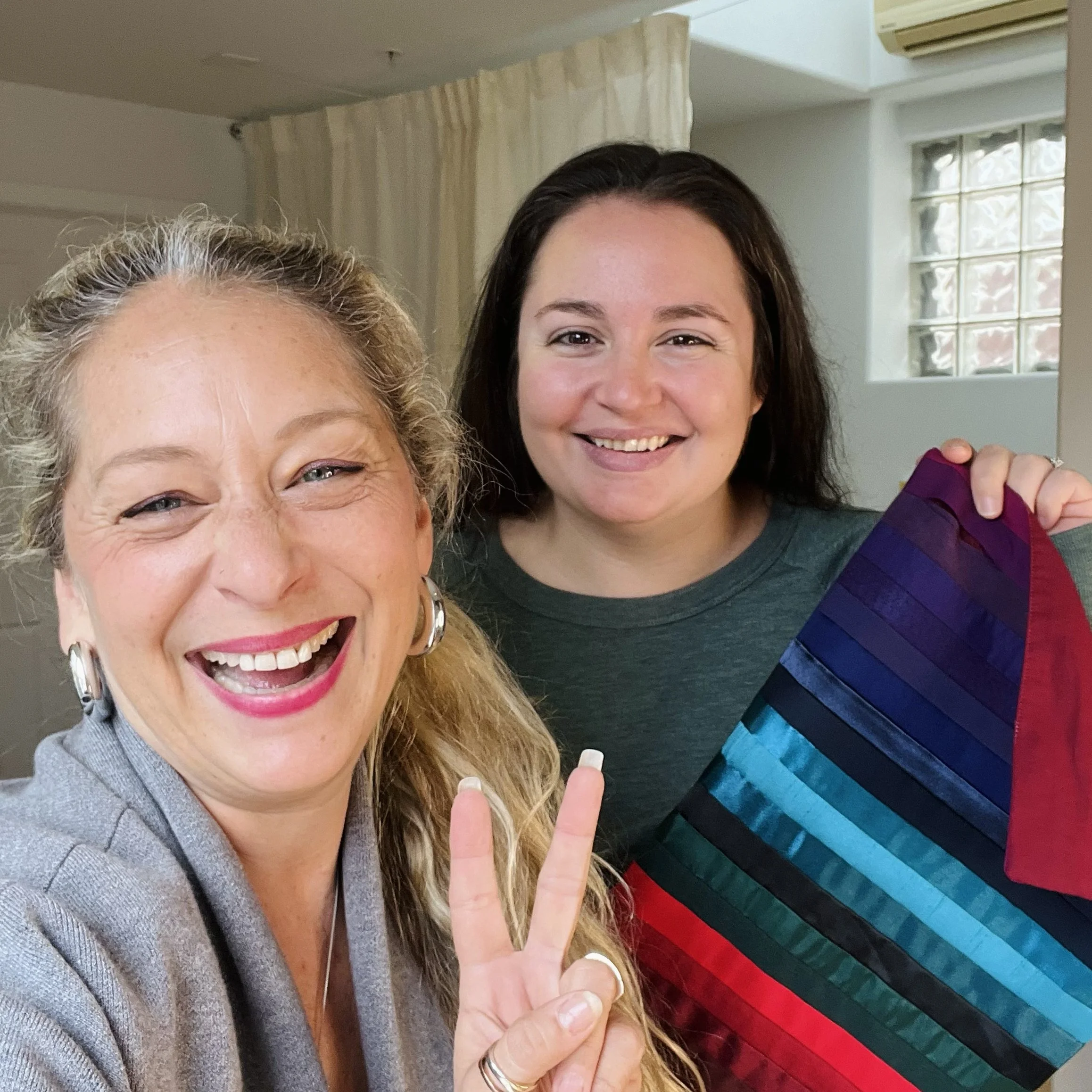 Two women smiling indoors, one holding a color sample or fabric with rainbow stripes.