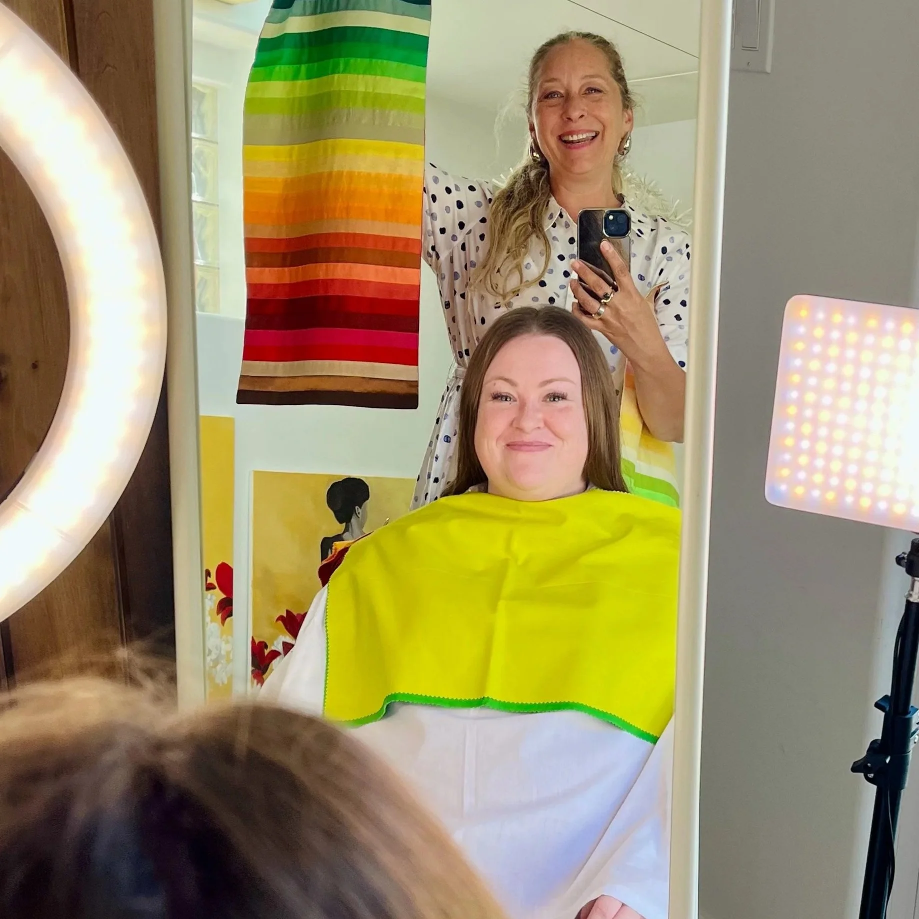 A woman with long brown hair sitting in a salon chair, wearing a yellow cape, while another woman takes a mirror selfie behind her, both smiling.