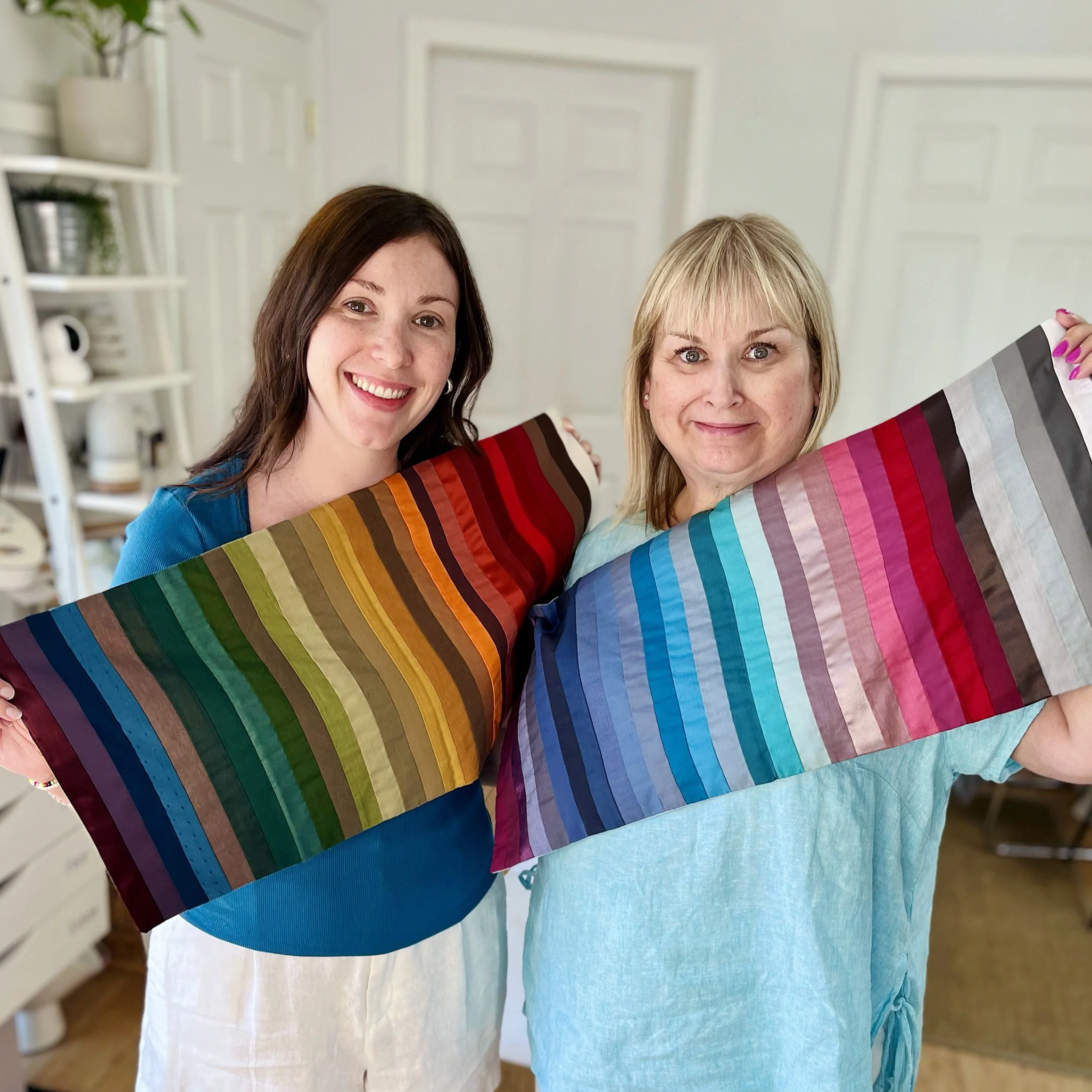 Two women smiling and holding colorful striped fabric panels in a bright room.