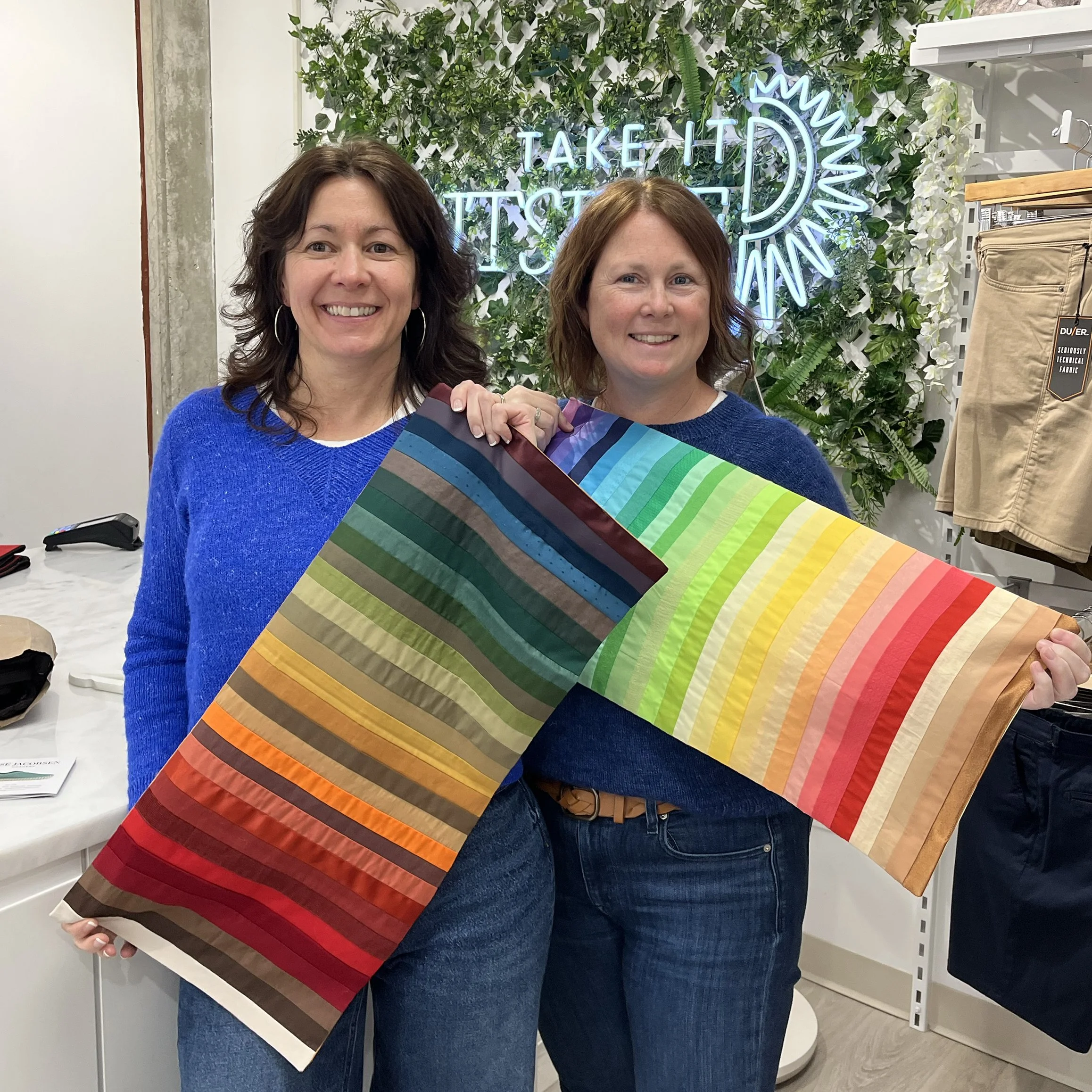 Two women smiling and holding a rainbow-colored fabric with stripes of various colors from dark blue to red. They are standing inside a store with a neon sign in the background that reads, 'Take It To The Next D.' The store has green foliage decor on the wall behind them.