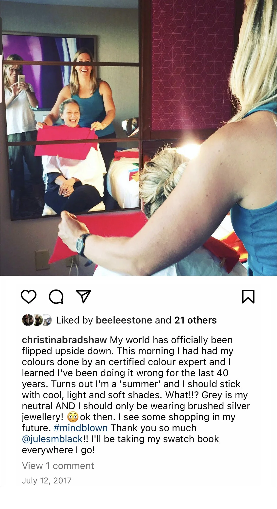 A woman is looking into a mirror while another woman stands behind her, holding a red paper or fabric, with two other women visible in the reflection of the mirror, one taking a photo with a smartphone.