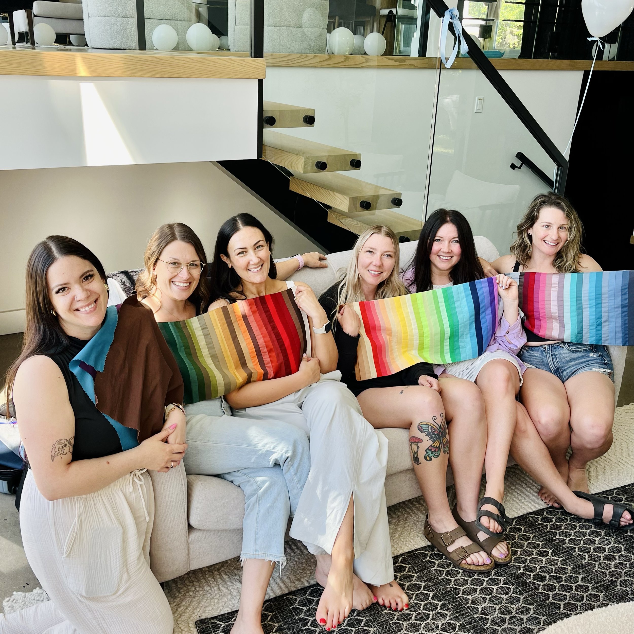 Six women sitting on a beige sofa, smiling and holding colorful rainbow patterned fabric, in a modern indoor setting.