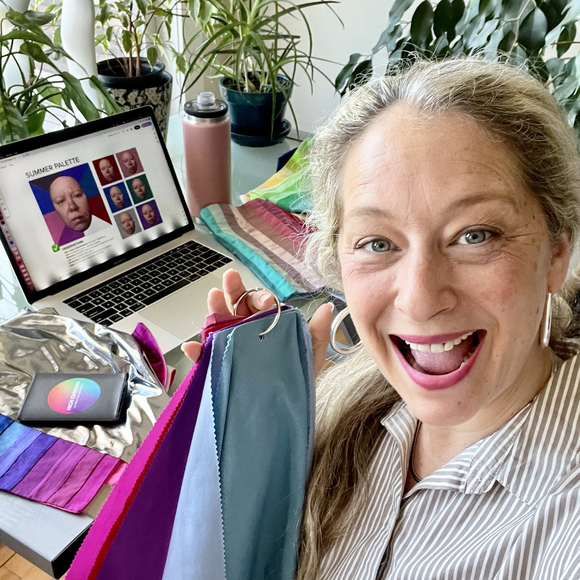 A woman with long blonde hair smiling and holding fabric swatches in front of her, with a colorful rainbow-striped pillow, a laptop displaying a color palette, and green plants in the background.