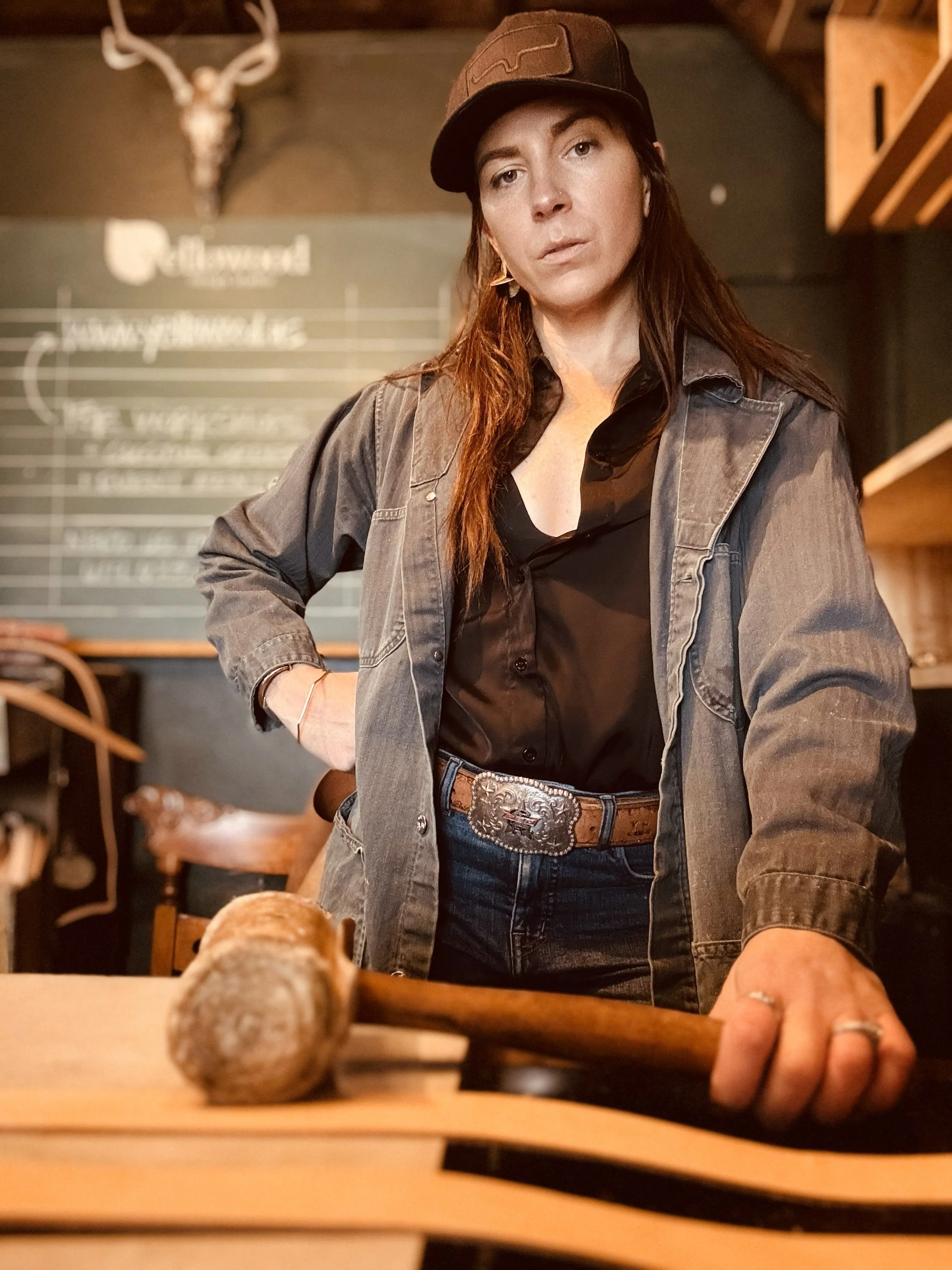 A woman with long brown hair wearing a black shirt, gray jacket, and a brown cap, stands in a rustic room with a wood-paneled wall, a blackboard with writing, and a mounted deer skull. She has a serious expression, hands on a wooden table with a sledgehammer, and is wearing rings and a leather belt.
