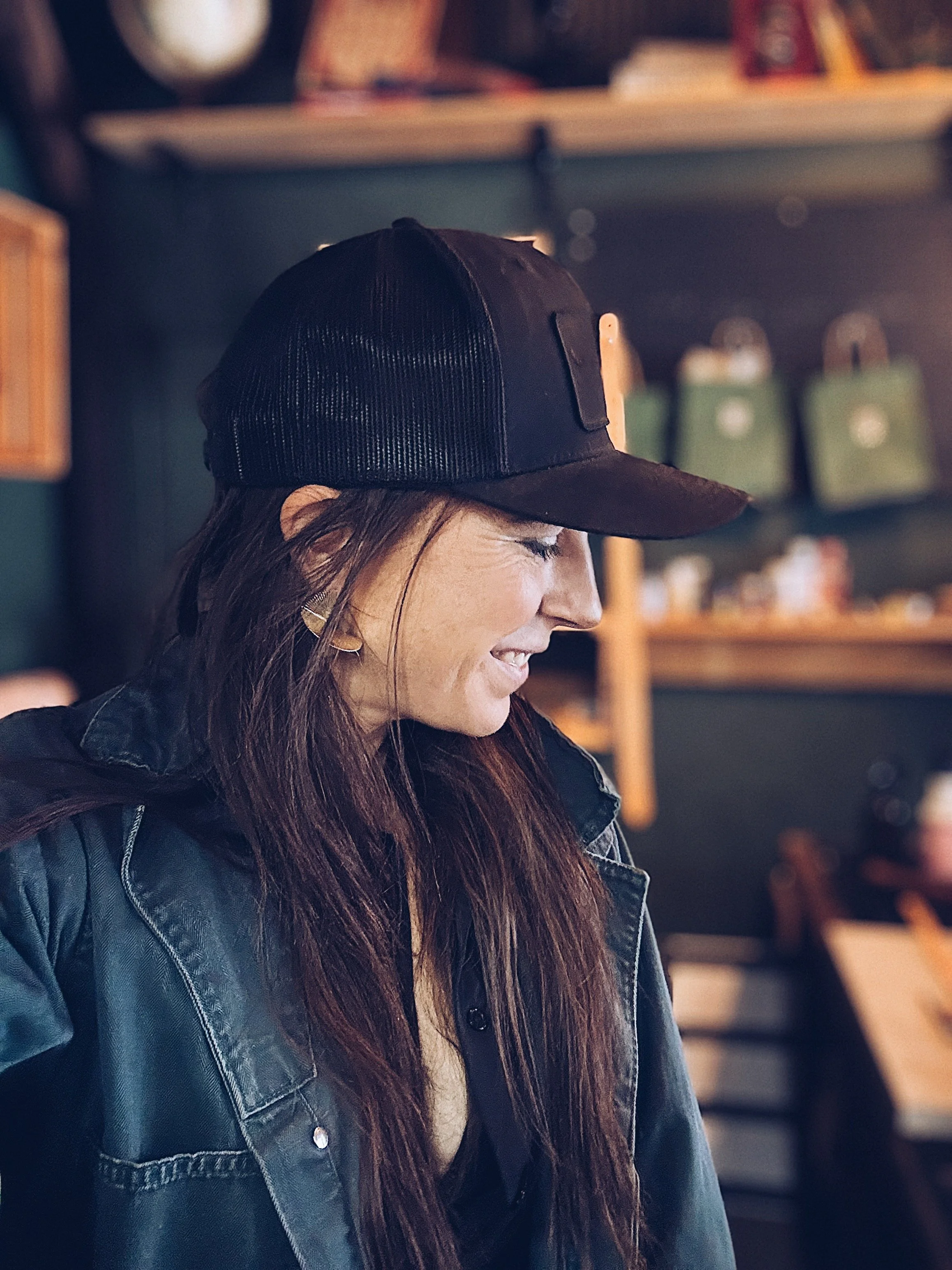 A woman with long brown hair wearing a black cap and black leather jacket, smiling and looking to her right indoors with shelves and green storage boxes in the background.