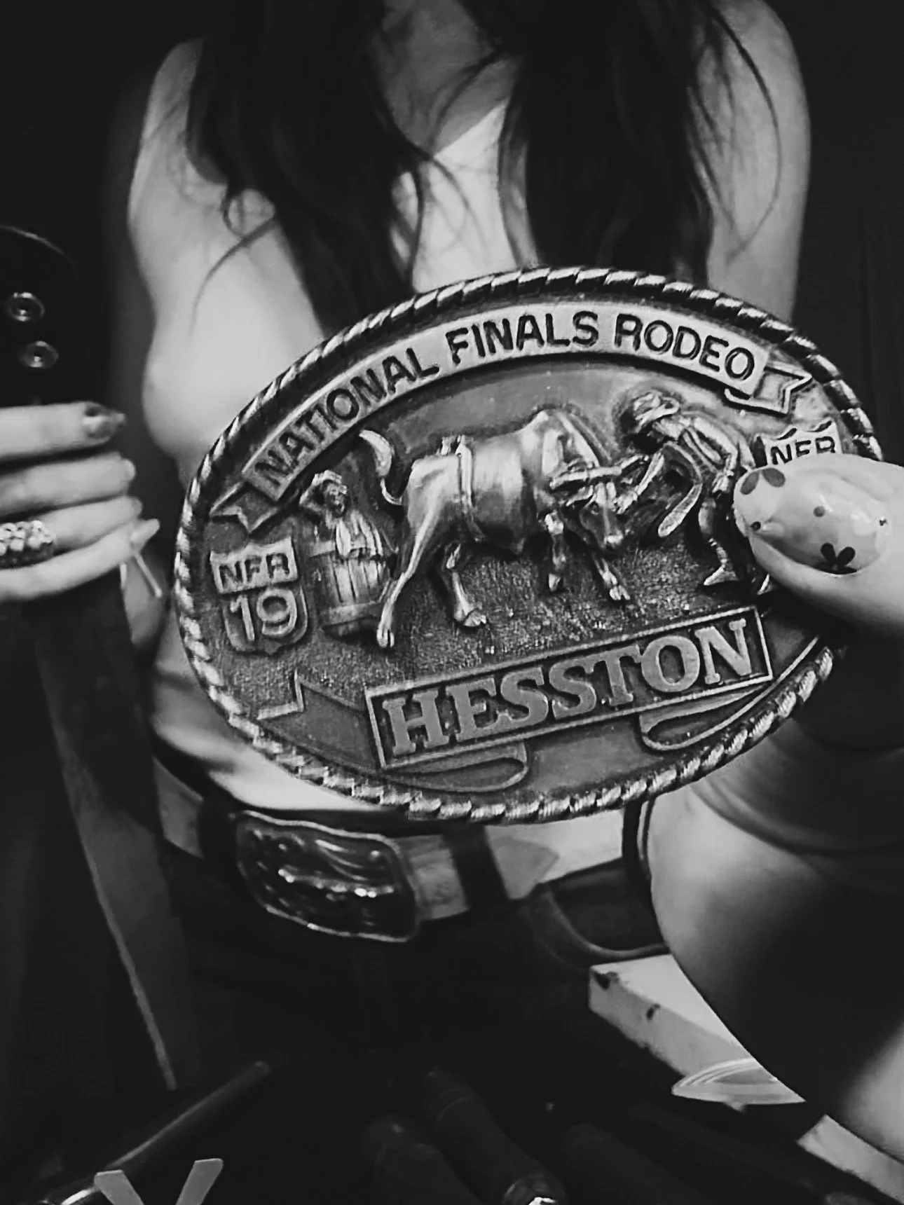 Black and white photograph of a woman holding a belt buckle or badge that reads 'National Finals Rodeo 19 Hessison,' featuring a cowboy riding a bucking horse.