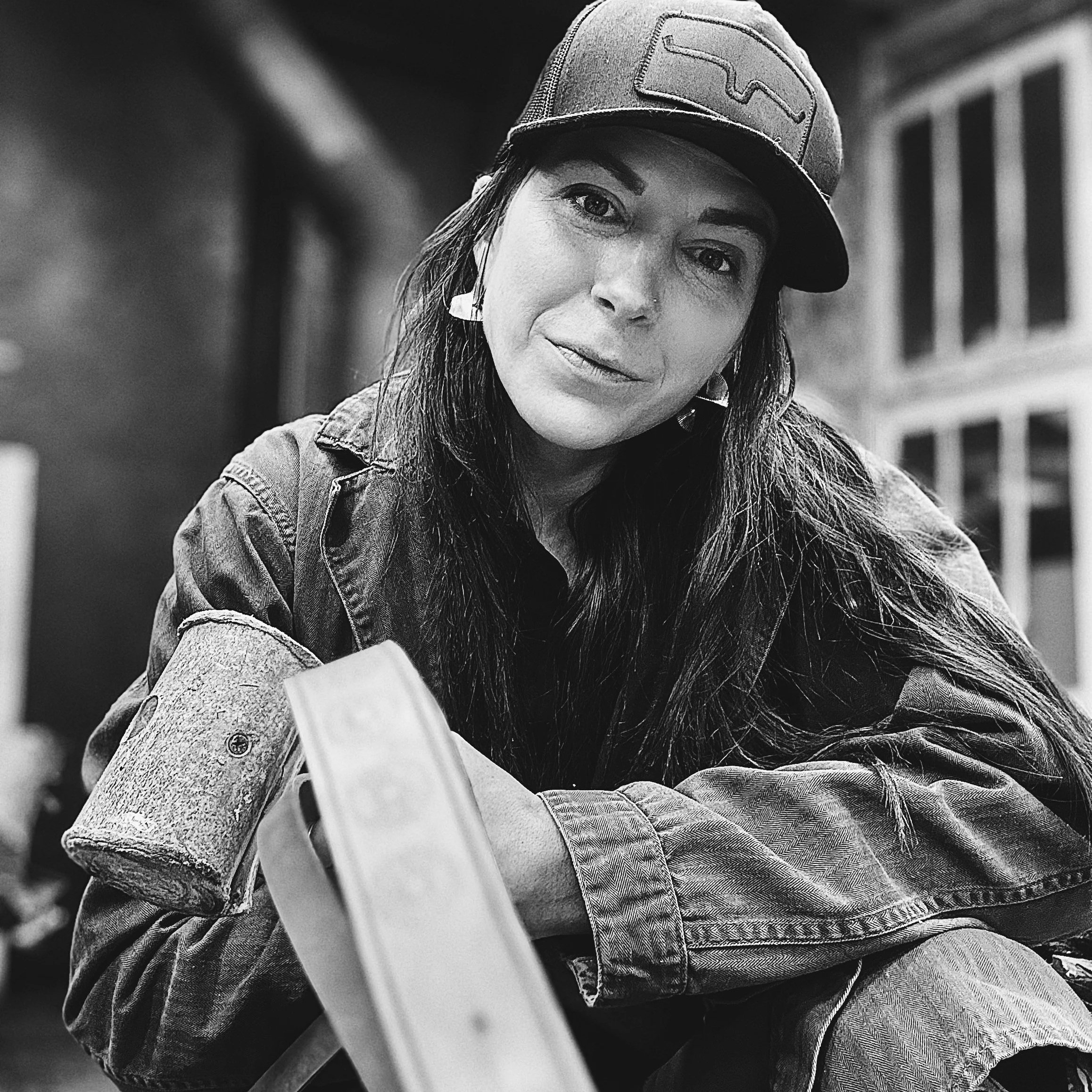 Black and white photo of a woman with long dark hair, wearing a baseball cap and denim jacket, sitting outdoors near a building with large windows.