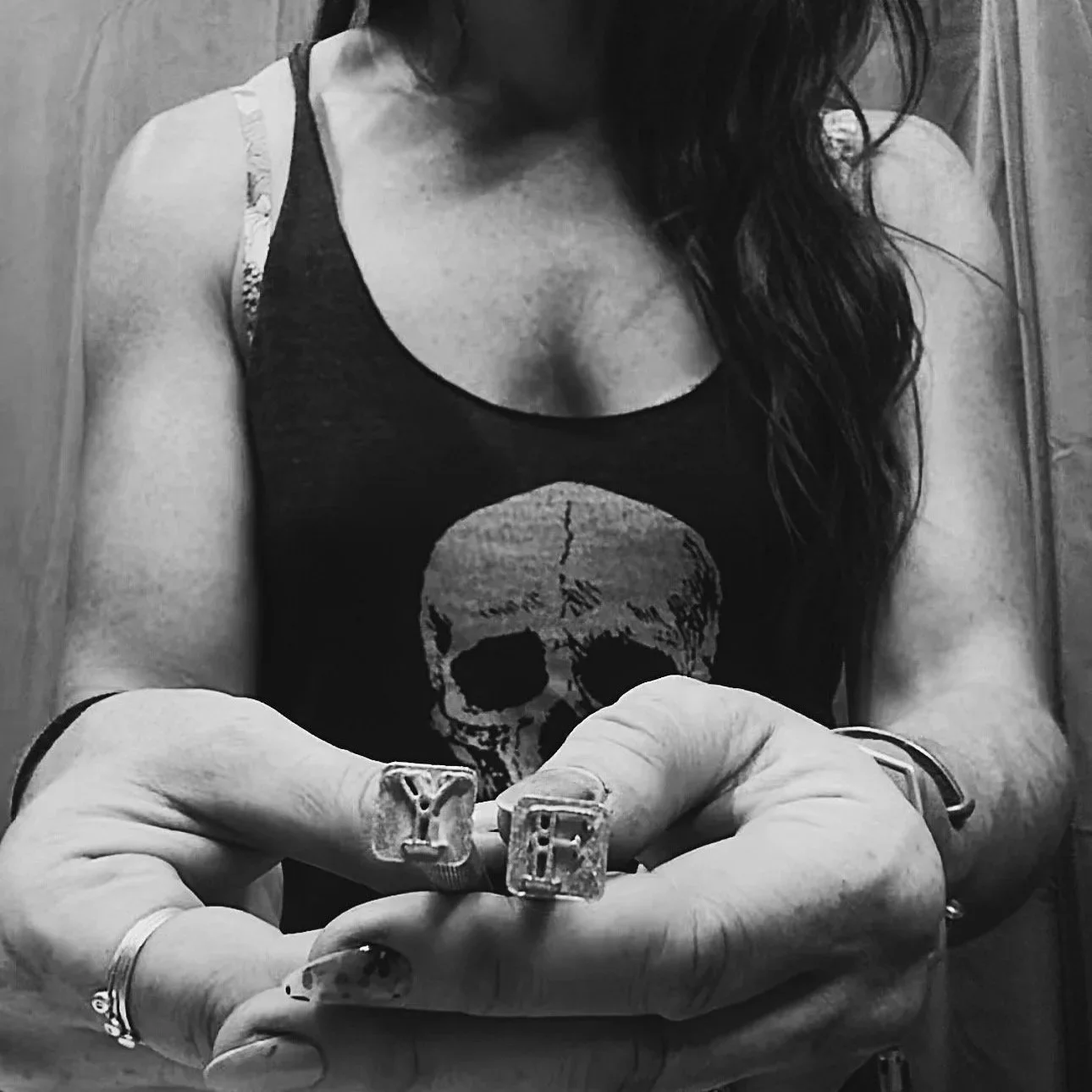 A woman with long hair wearing a tank top with a skull design, holding her hand with rings and decorated nails, in a grayscale photo.