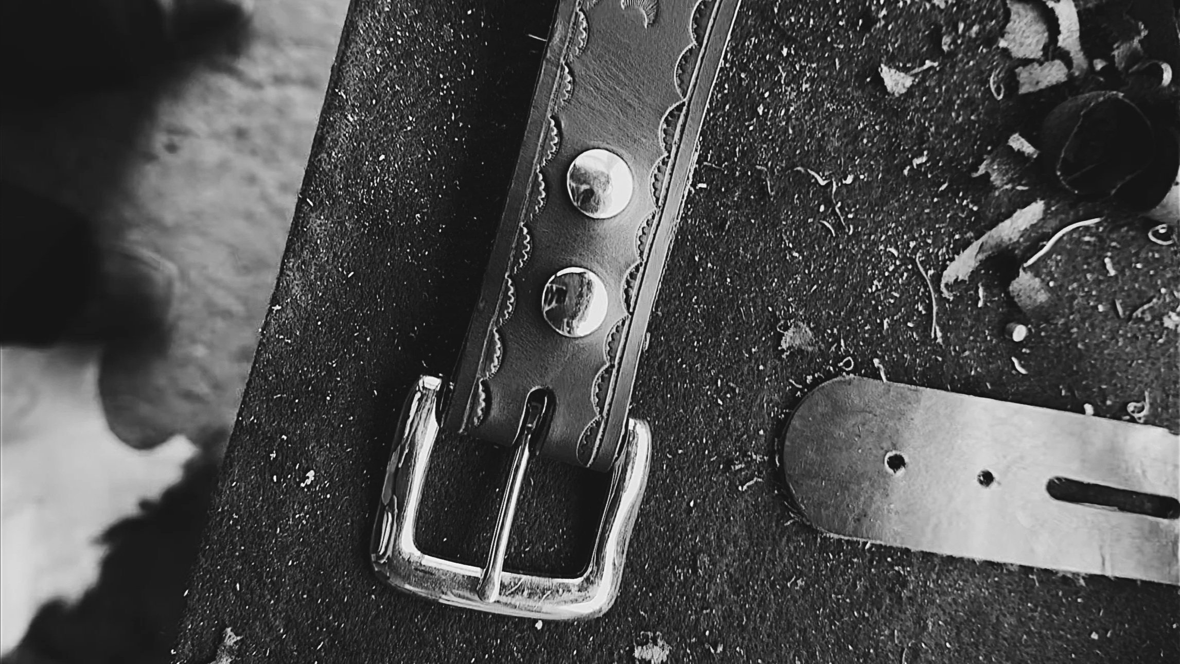 Black and white close-up of a belt buckle, a leather strap, and a metal tool on a rough surface with dirt and small debris.