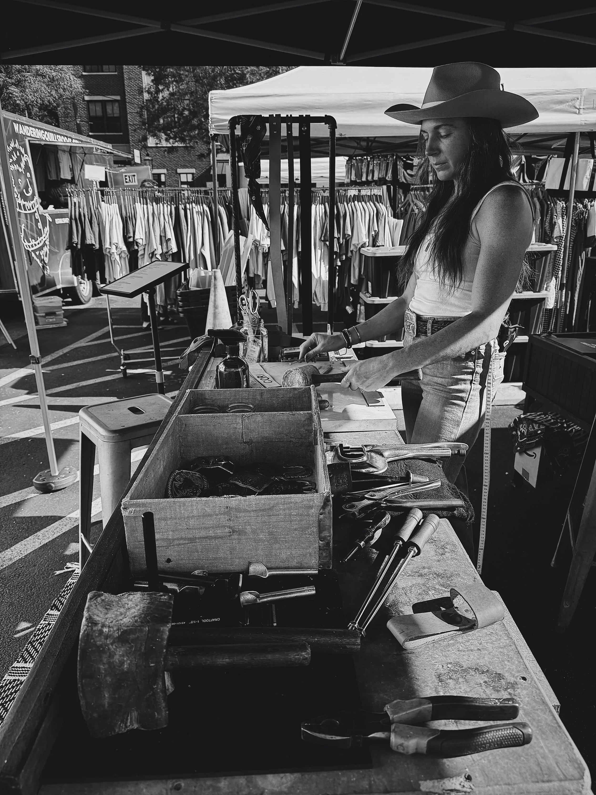 A woman wearing a wide-brimmed hat working at a booth with tools and craft supplies, selling clothing and accessories at an outdoor market.
