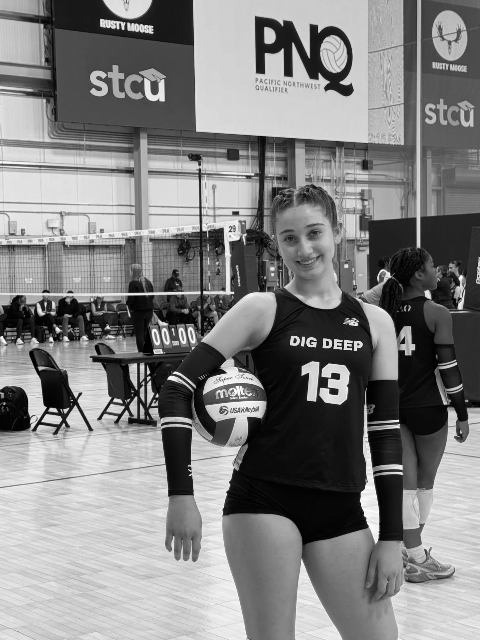 A young female volleyball player holding a volleyball on an indoor court, smiling at the camera. She is wearing a jersey with the number 13 and “Dig Deep” written on it. In the background, other players and people are visible, with banners and signs overhead.