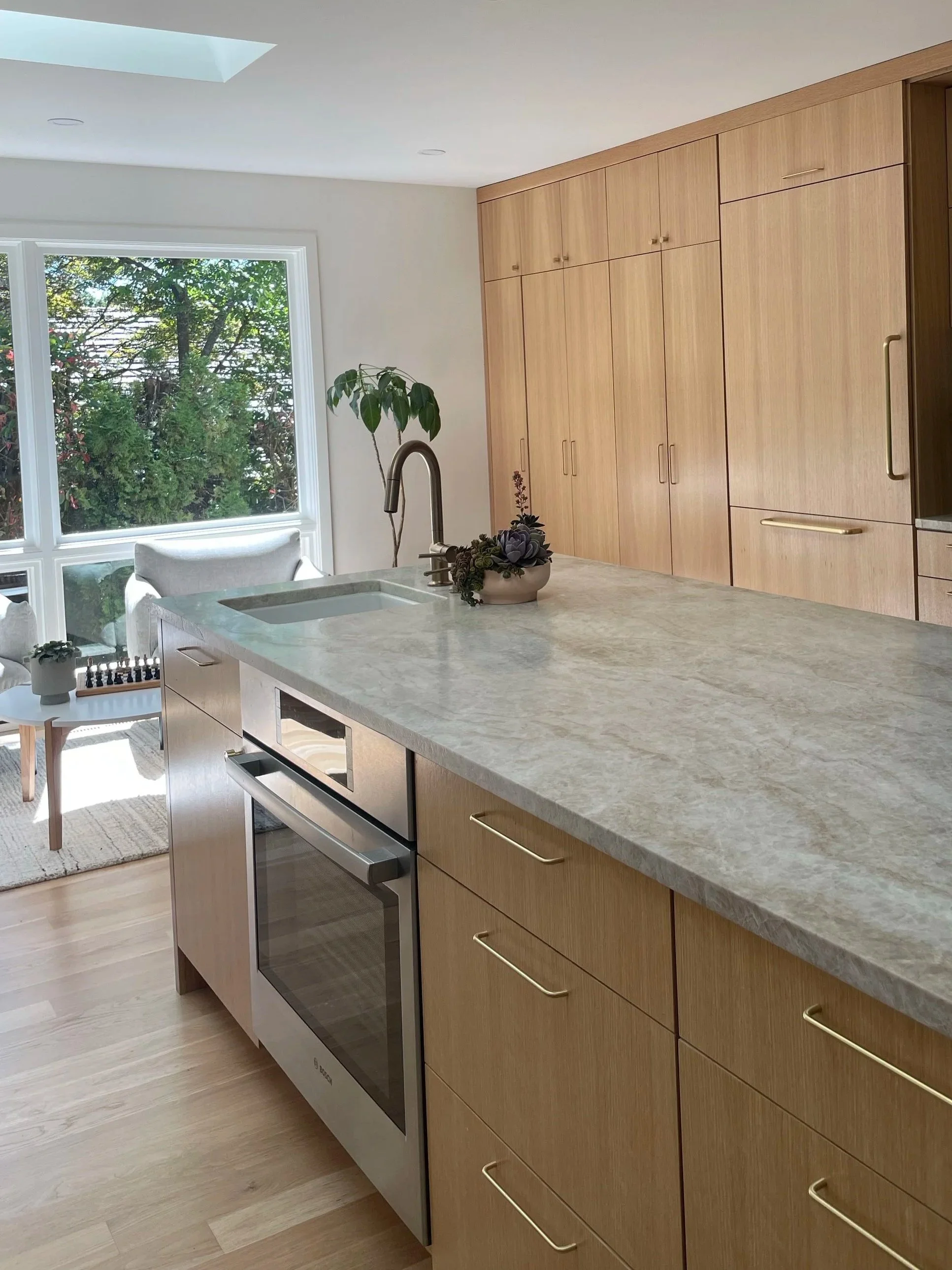 Modern kitchen with light wood cabinets, marble countertop, stainless steel oven, and large window with greenery outside.