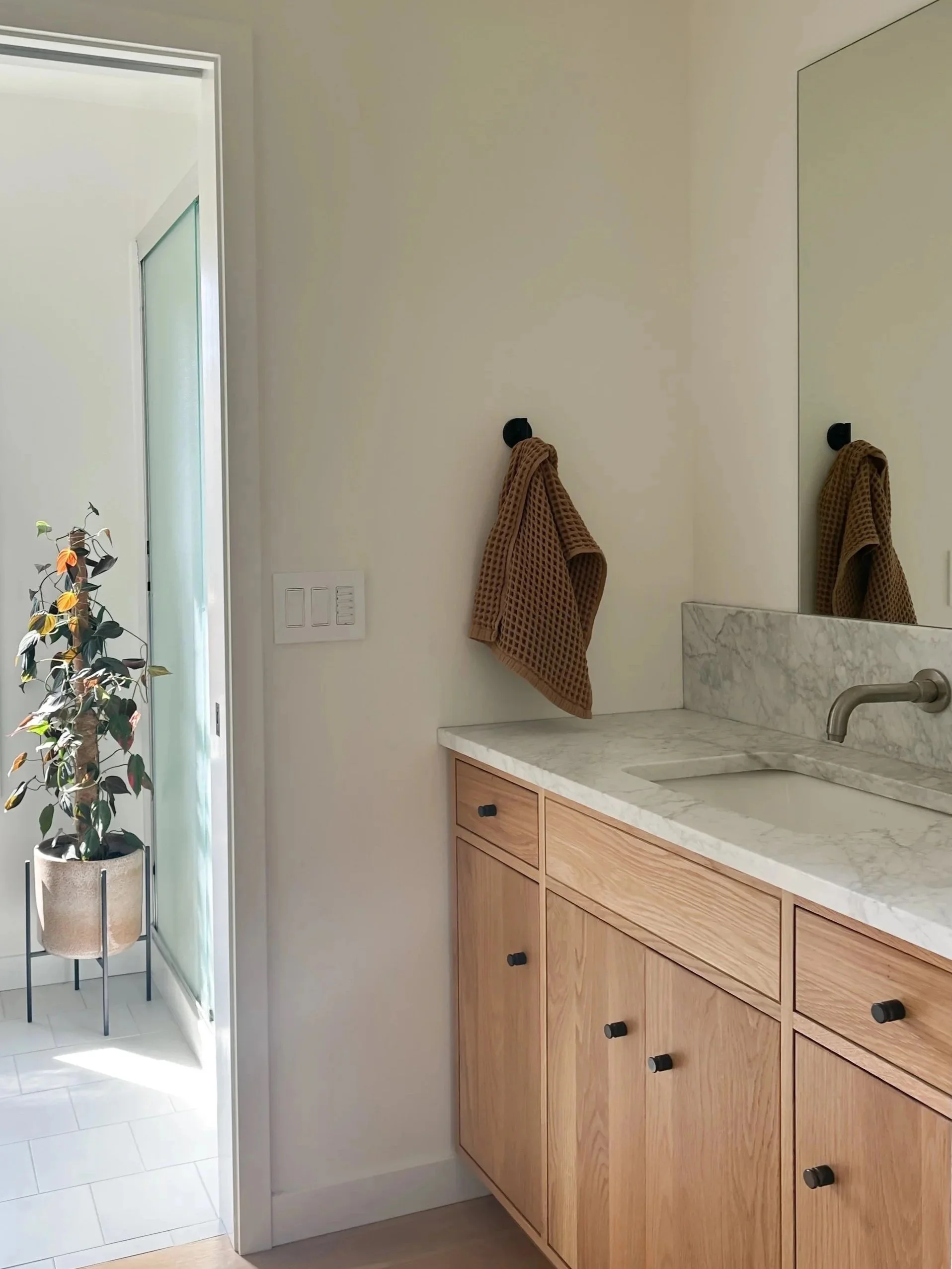 Bathroom with wooden cabinet, marble countertop, and a large mirror. A brown towel hangs on a black hook on the wall. Part of a door and a potted plant are visible in the background